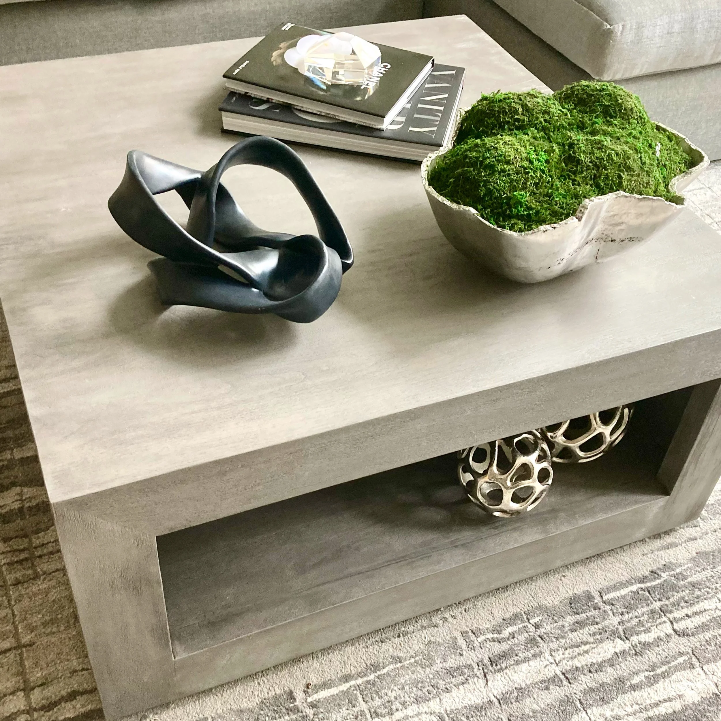 A modern coffee table with a stone top, on which are stacked magazines or books, a decorative glass object, and a large bowl filled with green moss or plants. Part of a gray sofa and a brick wall are visible in the background.