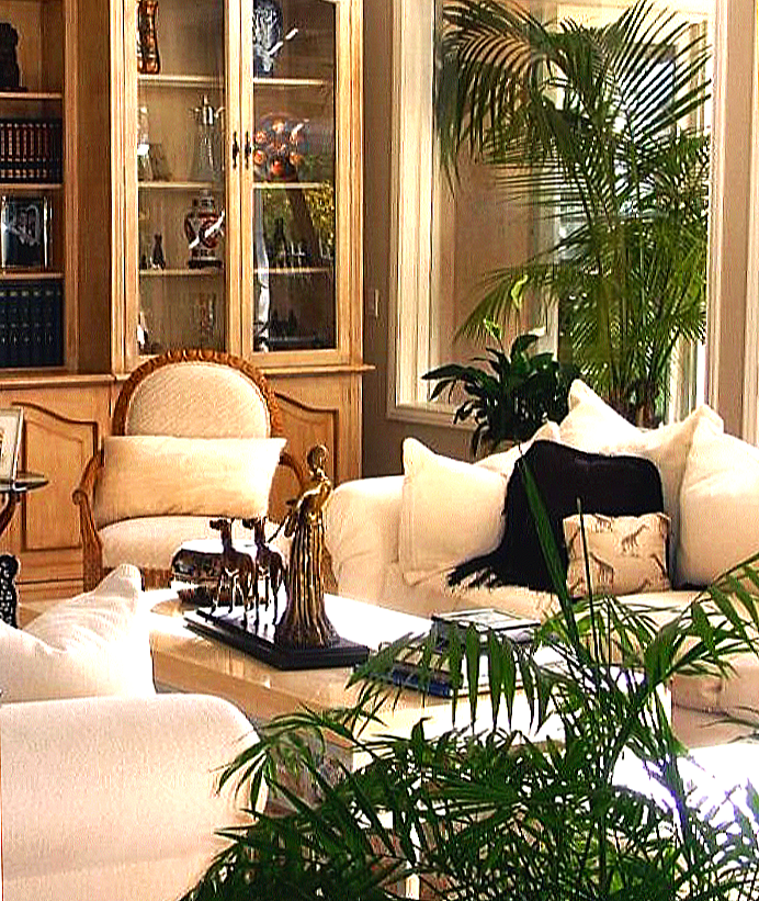 Living room with cream-colored sofa and armchair, glass-fronted wooden display cabinet, large potted plant, and decorative statues on a coffee table.