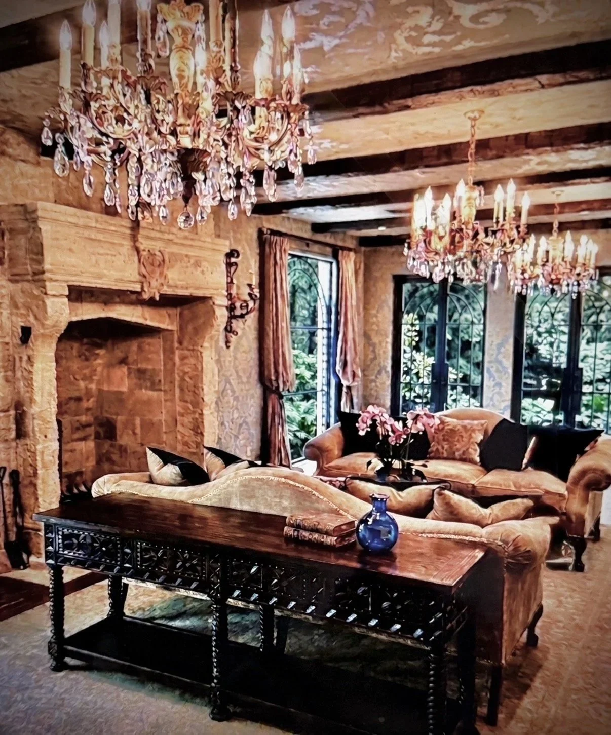 Elegant living room with a large stone fireplace, crystal chandeliers, velvet sofas with black and pink pillows, a dark wood coffee table with a blue vase and a rolled-up rug, and large arched windows with floral curtains.