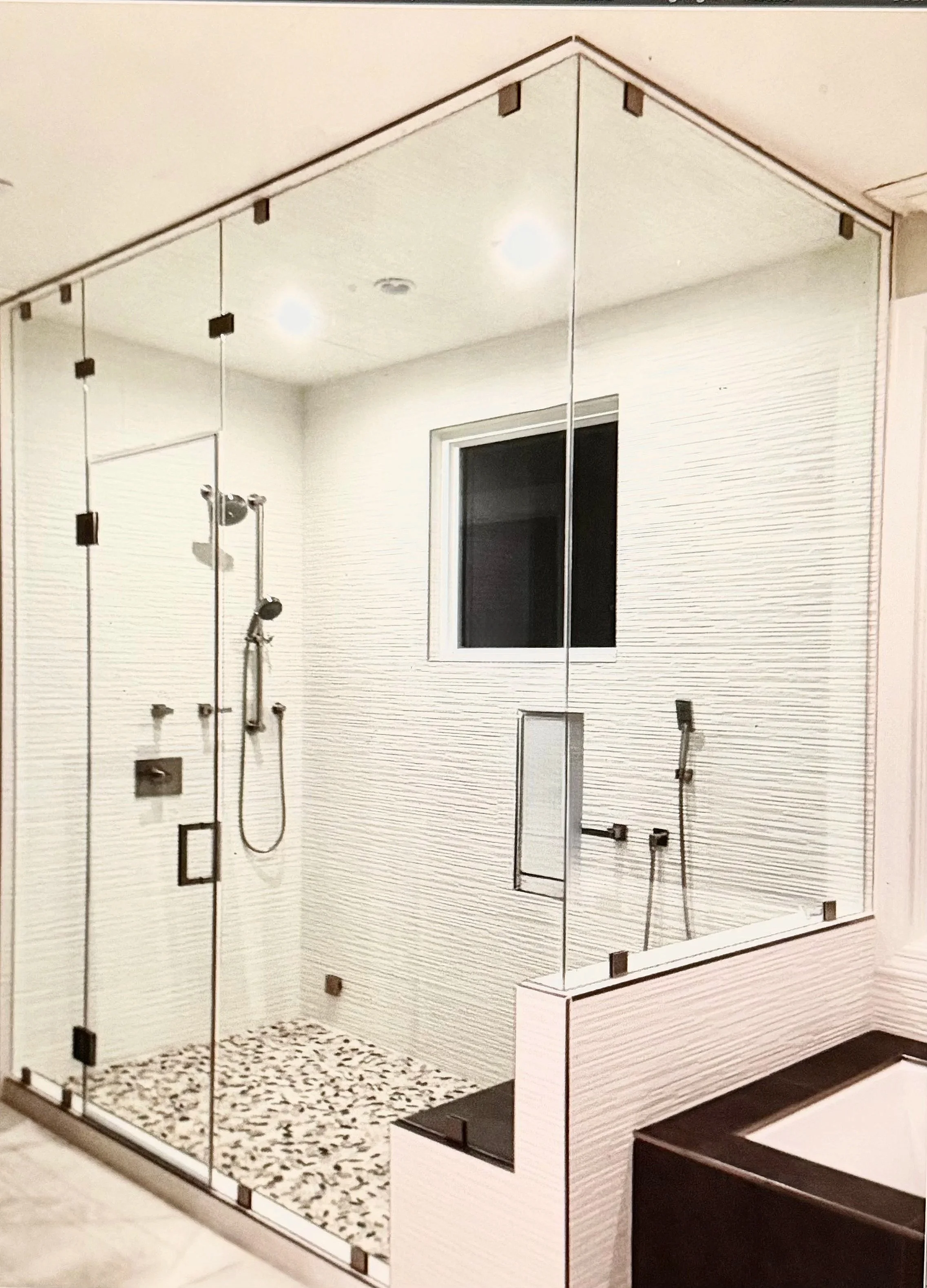 A modern glass shower enclosure with black metal framing and a bench, a wall-mounted shower head, and a window with white trim. The floor has a black and white pebble tile, and there is a white textured wall inside the shower.