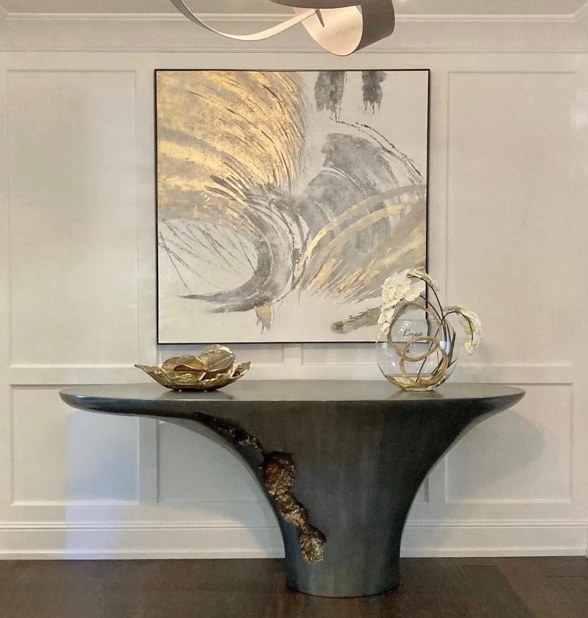A decorative console table with a curved black top and textured bronze-colored base, topped with a gold bowl and a clear glass vase containing white orchids, placed against a wall with a large abstract painting in shades of gray, black, and gold.