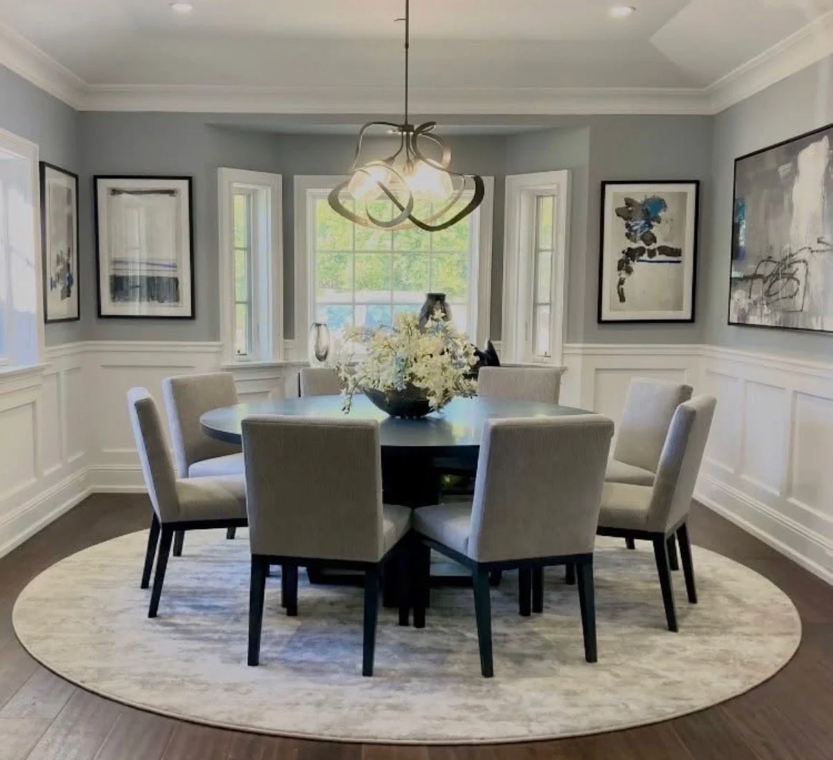 Dining room with a round table, eight cushioned chairs, a floral centerpiece, a modern chandelier, framed artwork on light gray walls, and large windows with a view of trees.