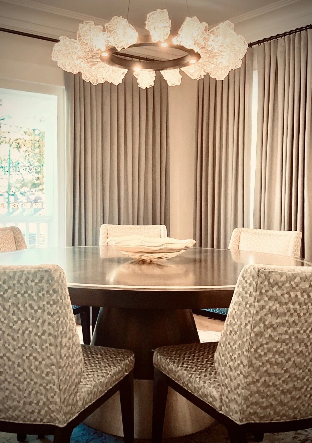 A dining room with a round table, five upholstered chairs, beige curtains, and a modern chandelier with cloud-like light fixtures.