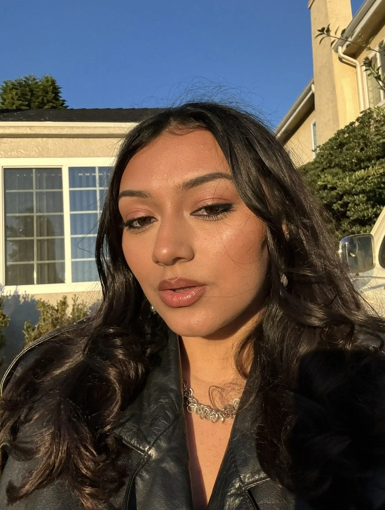 Close-up of a woman with long dark curly hair, wearing a black leather jacket and a silver chain necklace, outdoors in front of a house with a large window, against a clear blue sky.