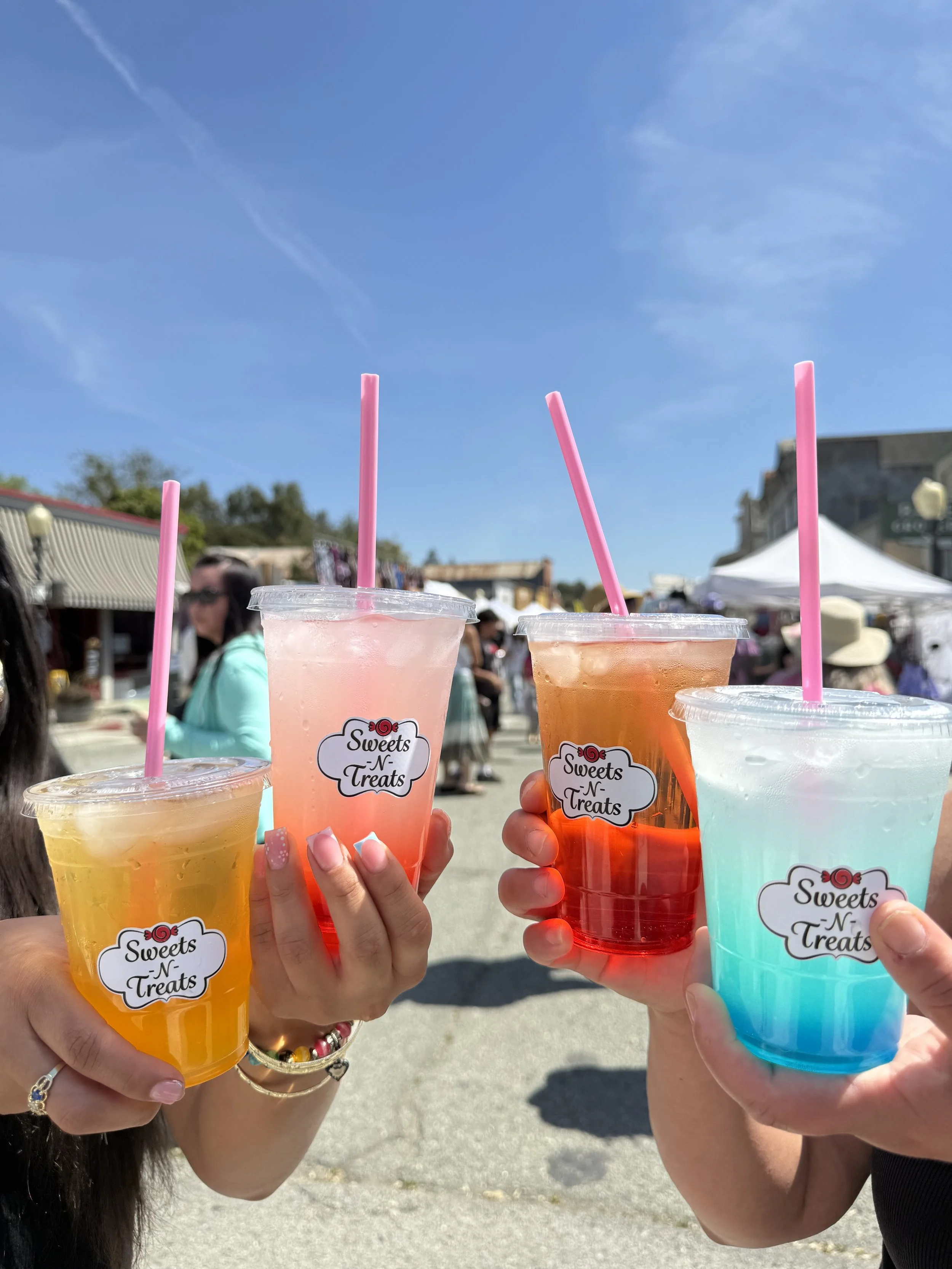Three people holding colorful drinks with pink straws at an outdoor event. The drinks are in clear cups with "Sweets N Treats" labels. The background shows a busy street with tents, buildings, and a clear blue sky.