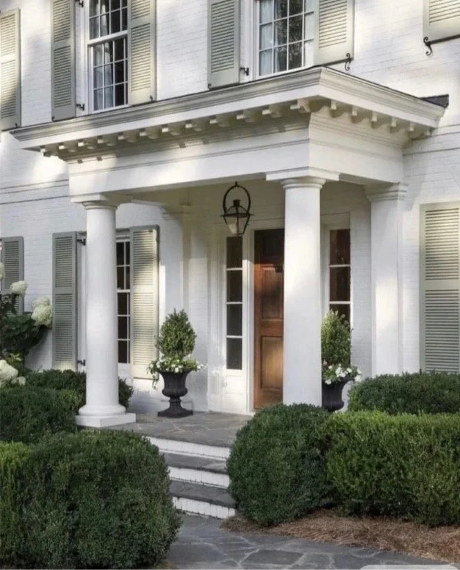 Front entrance of a white house with a covered porch supported by two large white columns. There are steps leading up to a wooden front door, flanked by potted plants and windows with shutters. The house has a classic, elegant appearance.