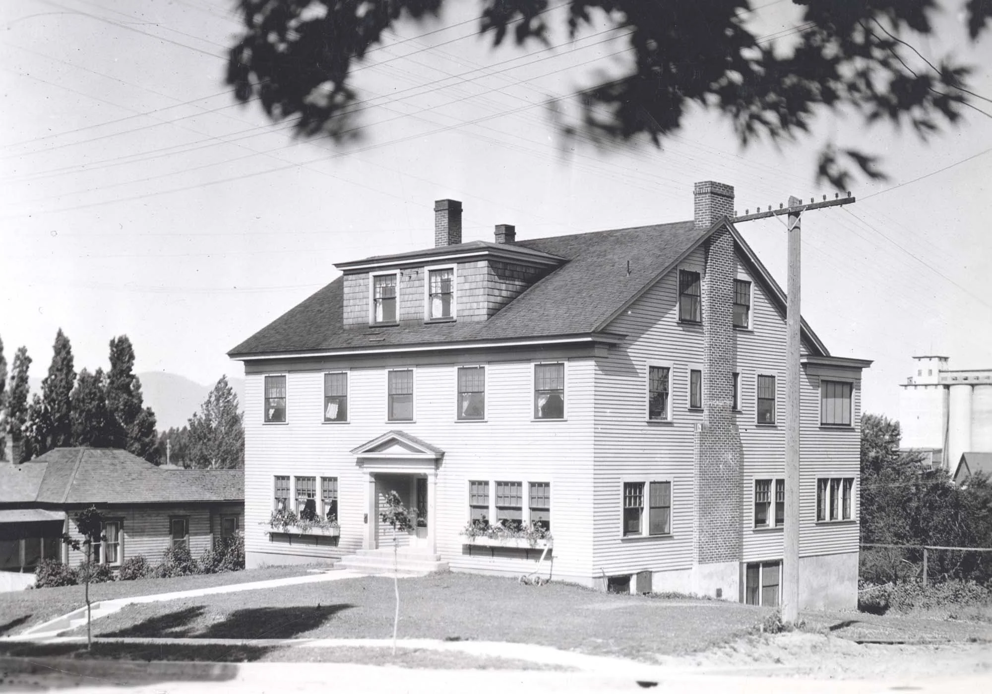 Black and white photo of a multi-story house with a gabled roof, chimney stacks, and a small porch at the front, surrounded by a lawn with a curved pathway, trees, and electrical wires overhead.