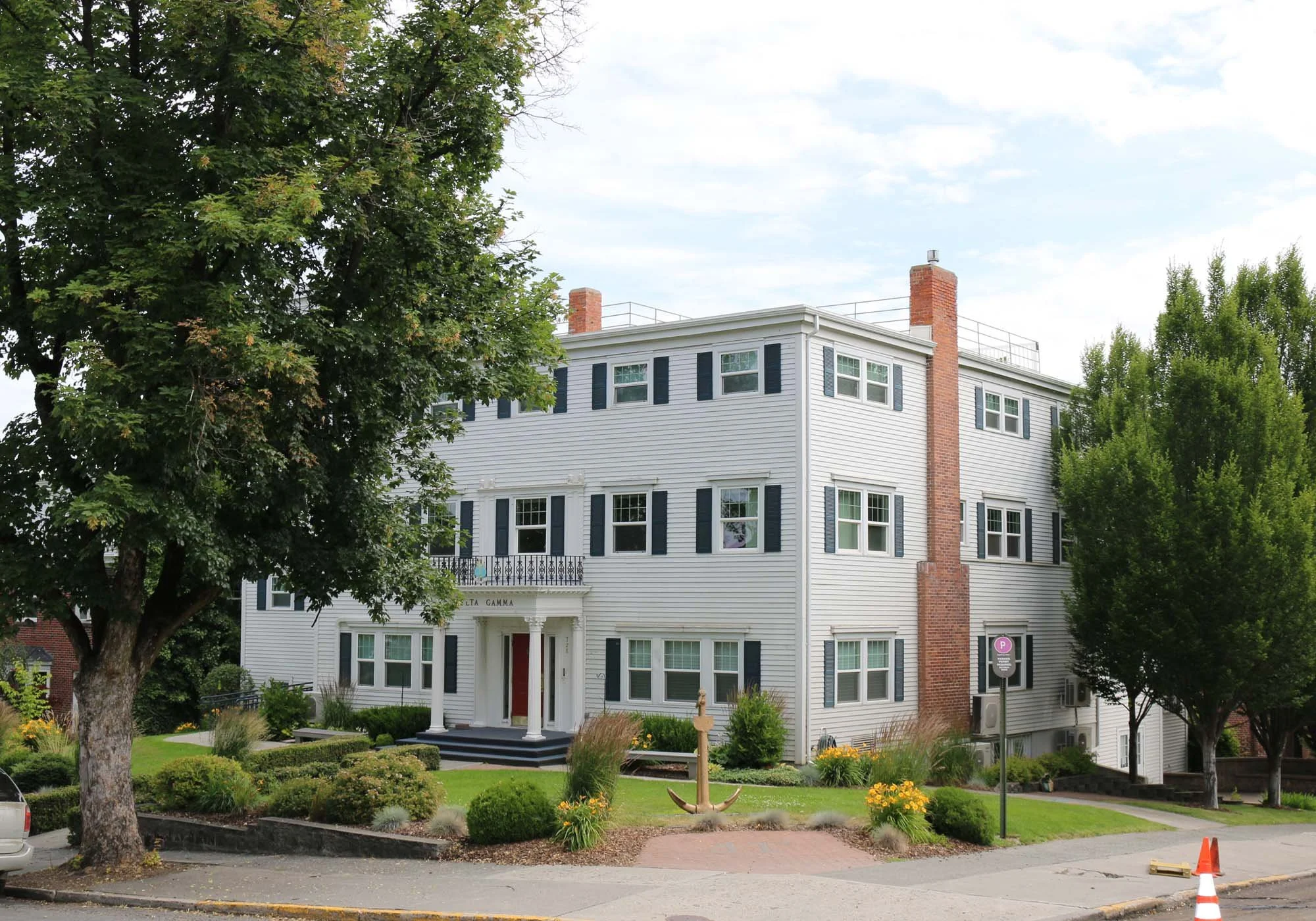 A white multi-story house with black shutters, a brick chimney, and a small front porch. The house is surrounded by a well-maintained garden with green shrubs and yellow flowers, trees on either side, and a sidewalk street in front.