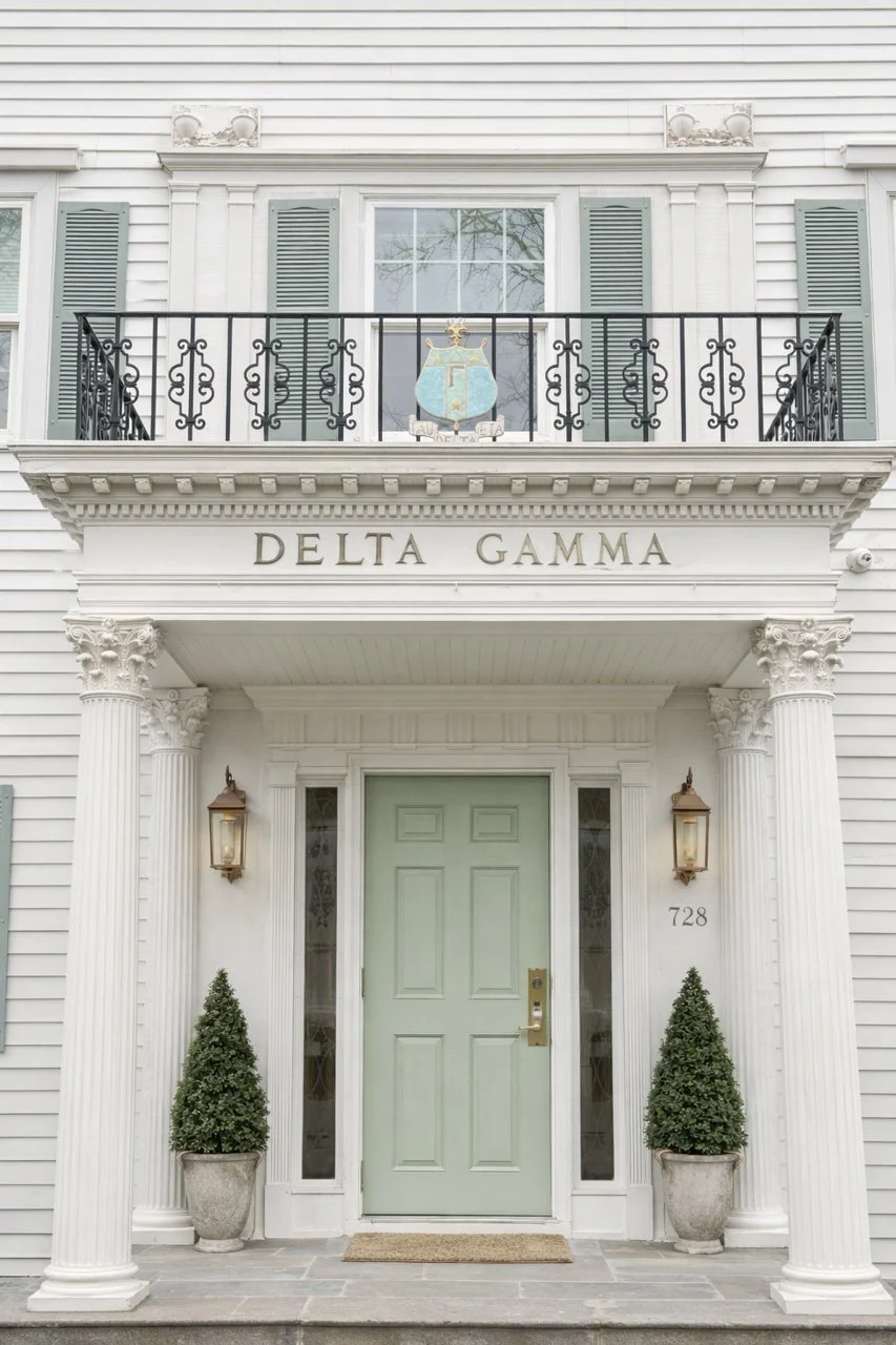Front entrance of a white house with green door, two potted shrubs, and classical columns, under a balcony with the Greek letters delta gamma and a crest.