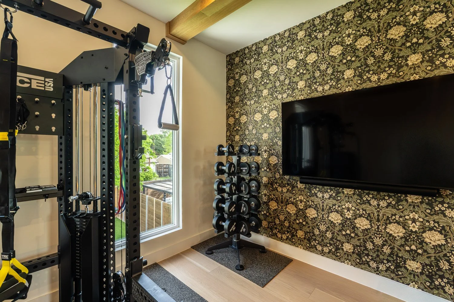 Modern home gym with Force USA functional trainer, Rogue dumbbell rack, wall-mounted TV, and natural light from window