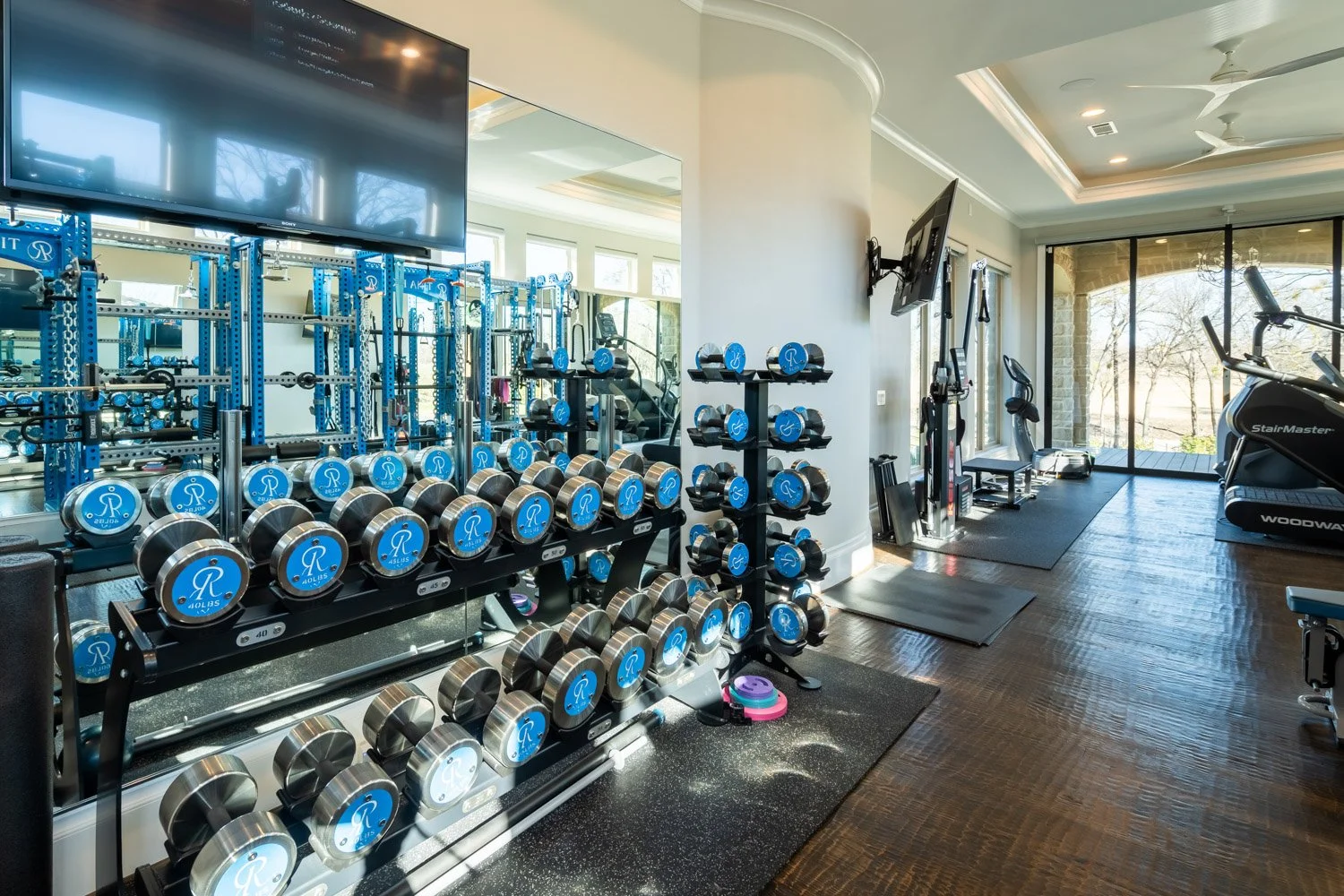 Luxury home gym with mirrored walls, organized dumbbell racks, blue custom strength equipment, and natural light