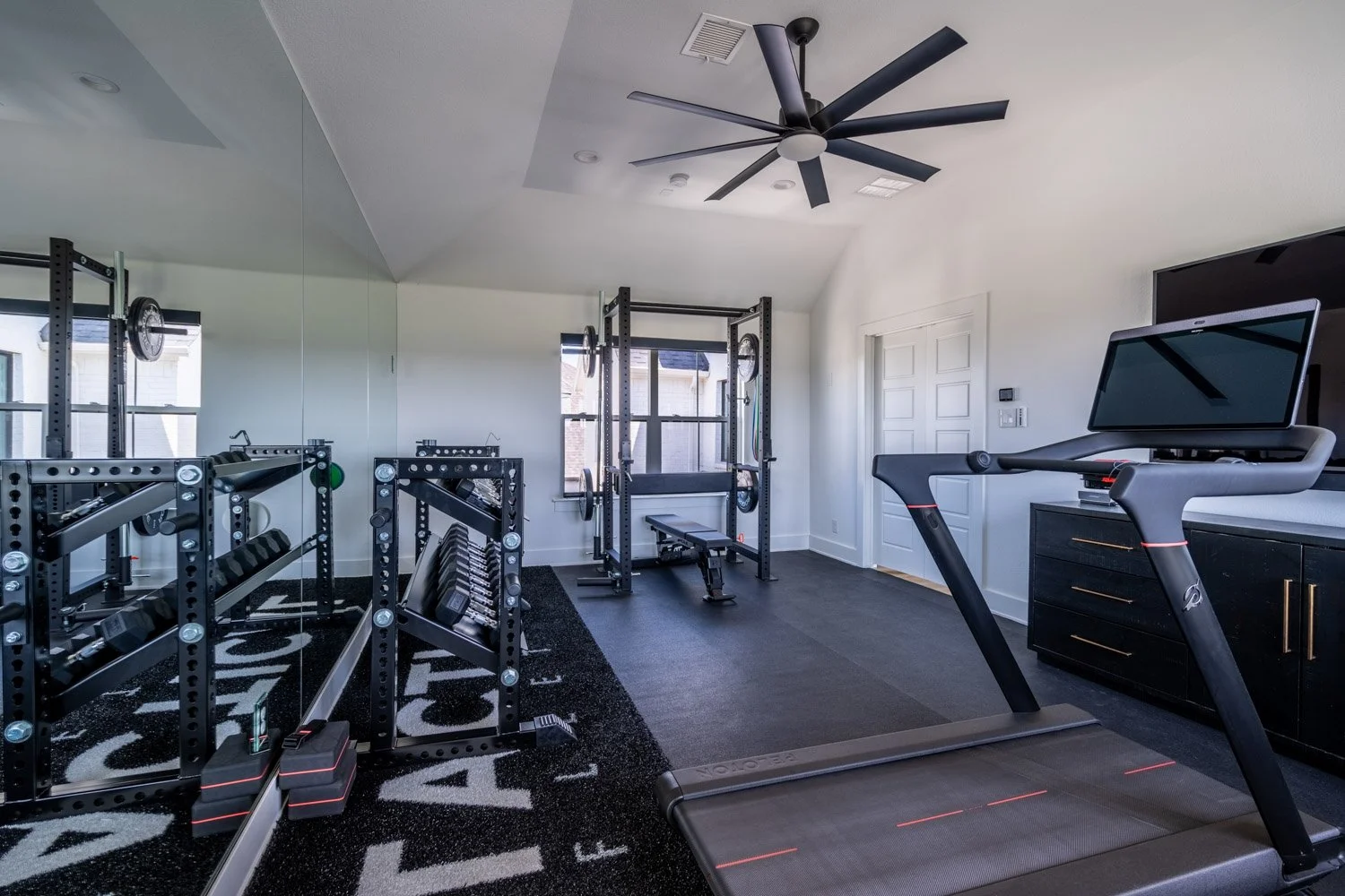 Modern custom home gym with treadmill, Rogue squat rack, dumbbell storage, mirrored wall, and high ceiling with ceiling fan
