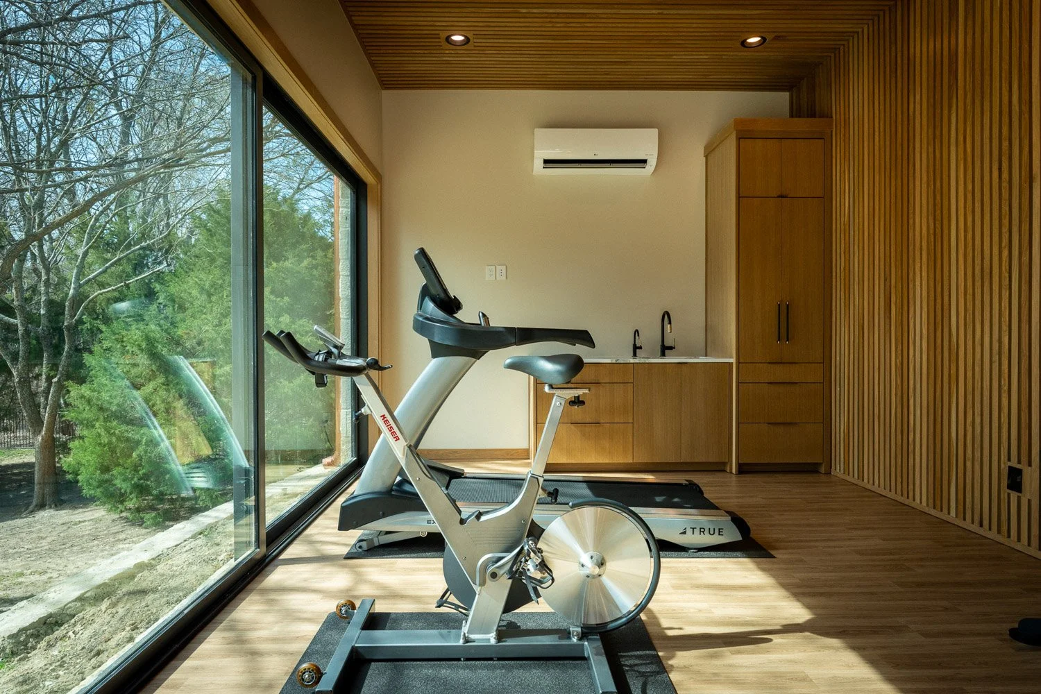 Modern custom home gym with stationary bike, treadmill, natural light, and warm wood paneling design
