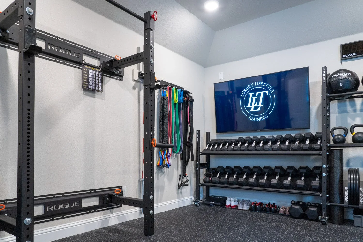 Custom home gym with Rogue squat rack, resistance bands, dumbbell storage, and wall-mounted TV display