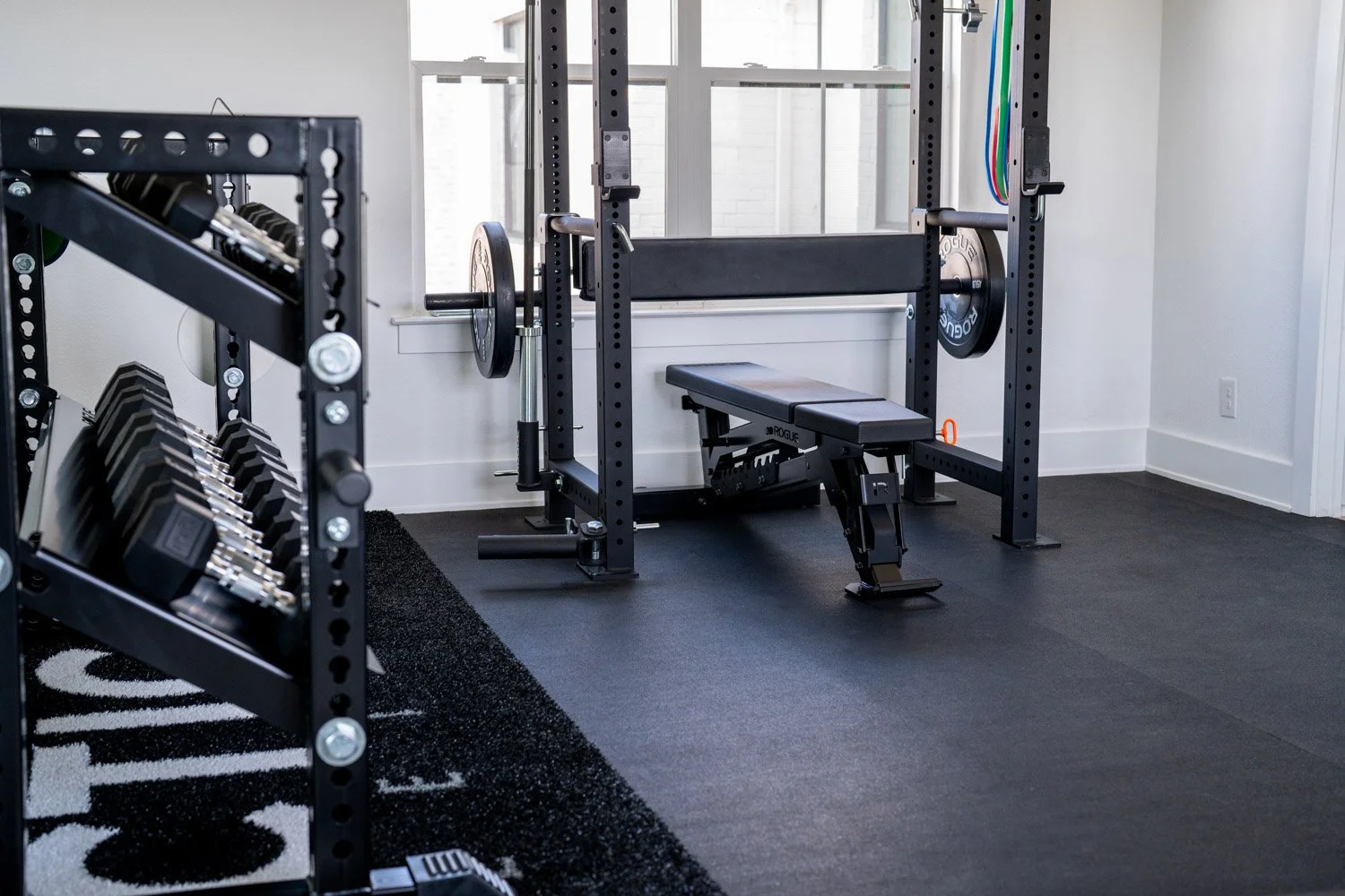 Custom home gym with Rogue dumbbell storage, squat rack, resistance bands, and clean modern design with natural light