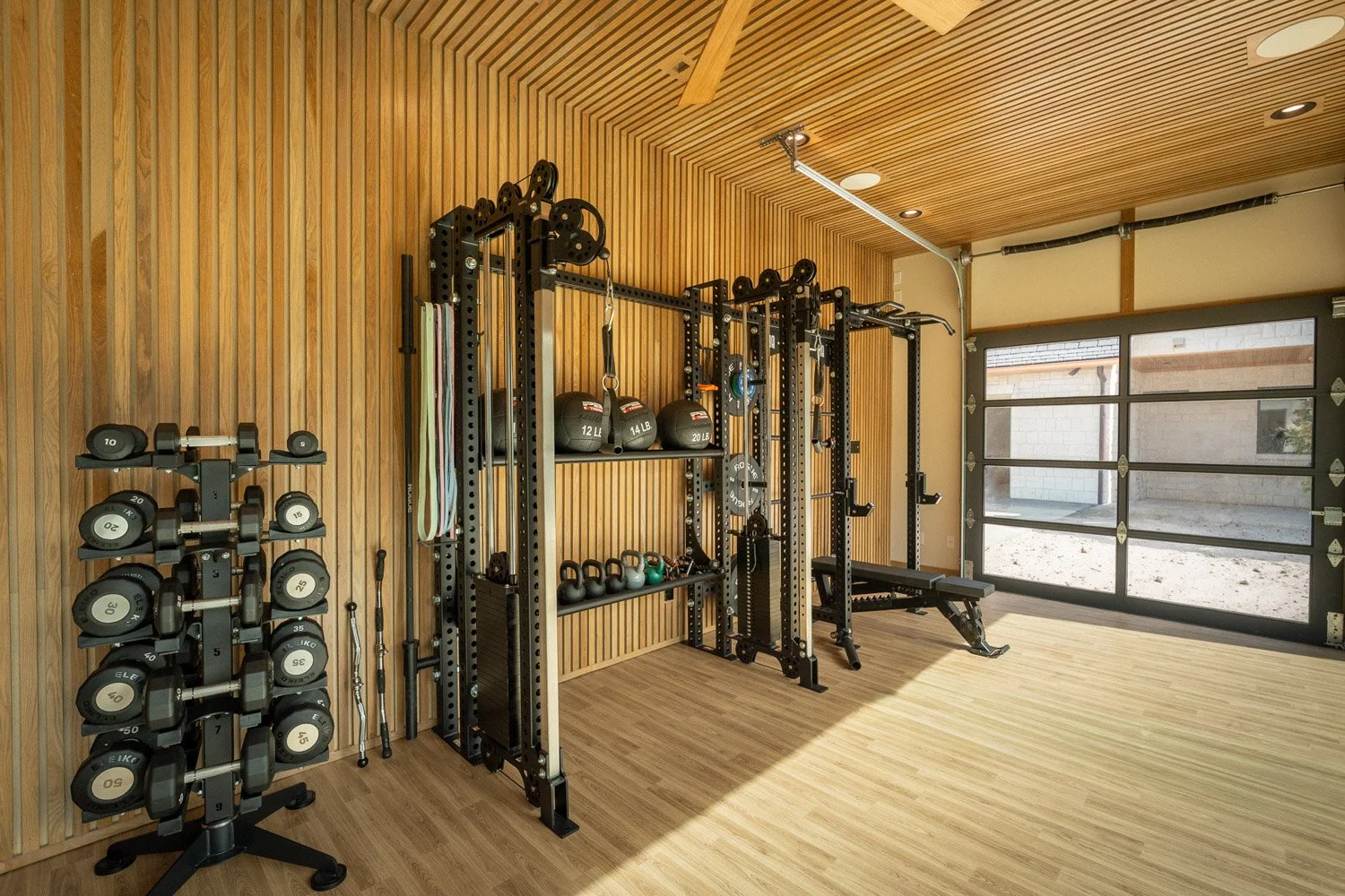Luxury custom home gym with dumbbell rack, cable machine, kettlebells, and wood slat walls with modern garage-style door