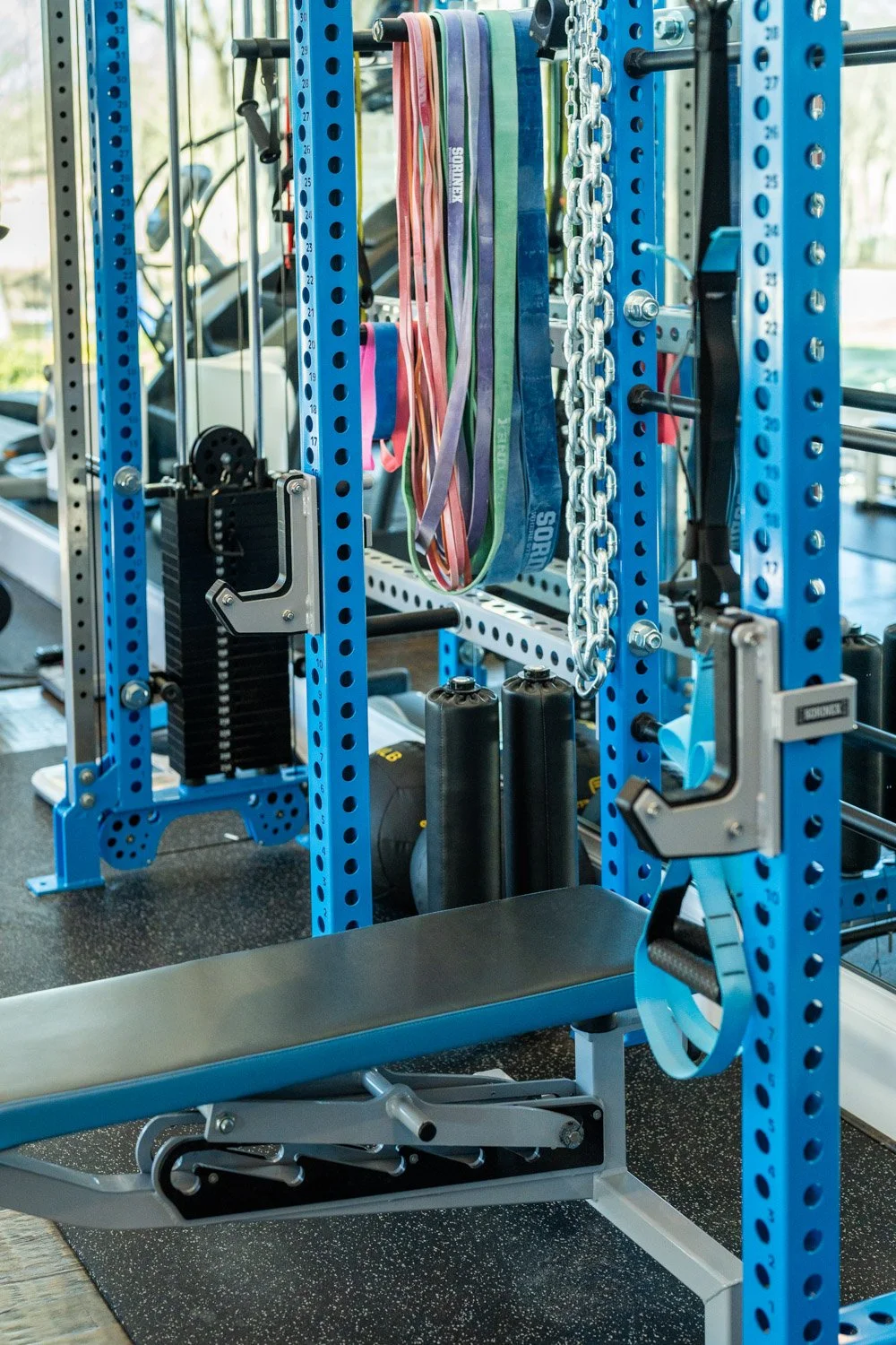 Custom home gym with blue strength rack, adjustable bench, resistance bands, chains, and cable machine system