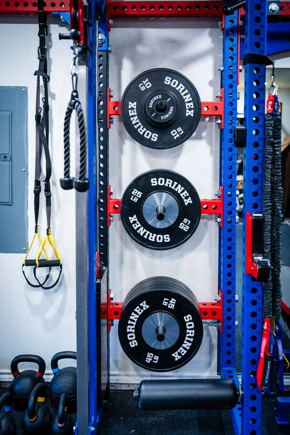 Close-up of Sorinex weight plates mounted on custom home gym rack with resistance straps and kettlebells below