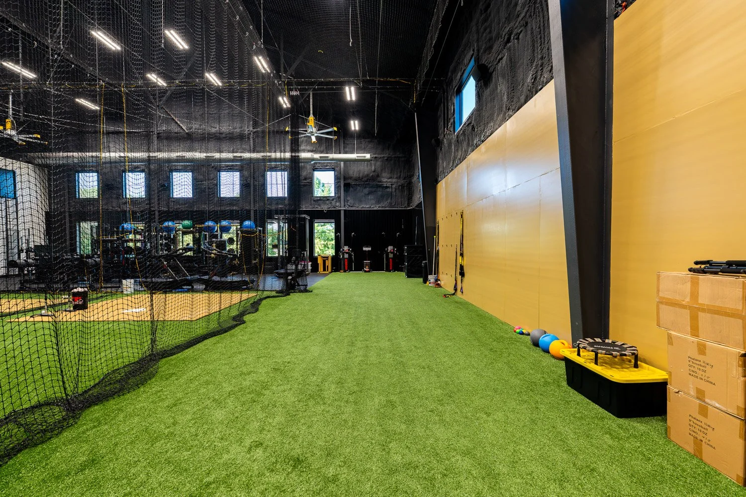 Indoor turf training lane in professional gym with netted batting cage, conditioning equipment, and open workout space