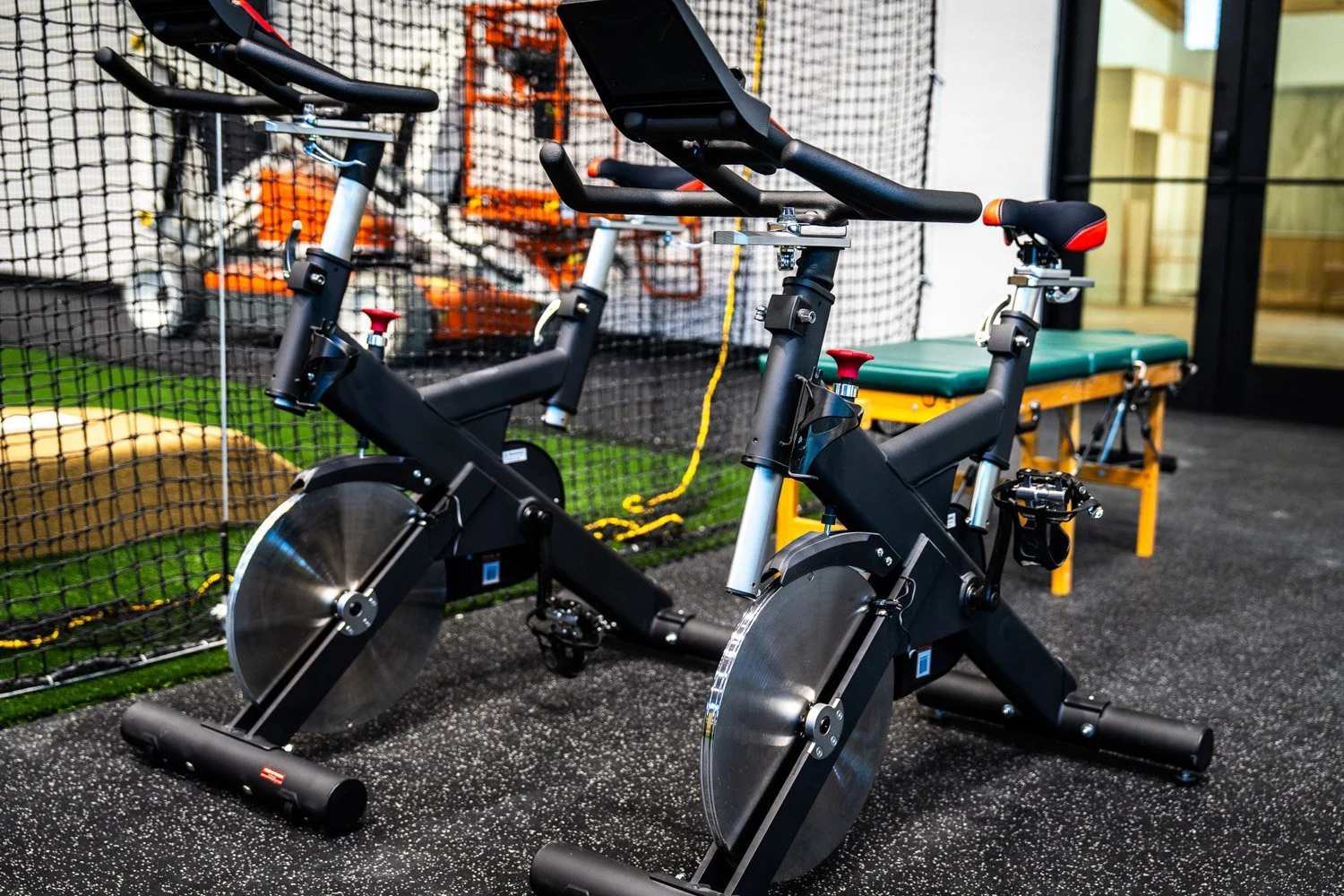 Indoor cycling bikes in custom training gym with turf flooring, batting cage netting, and performance workout area
