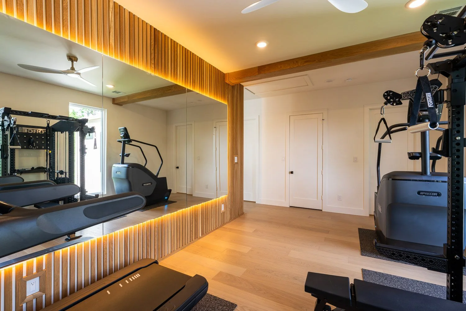 Modern custom home gym with treadmill, strength equipment, mirrored wall, wood slat paneling, and warm ambient lighting