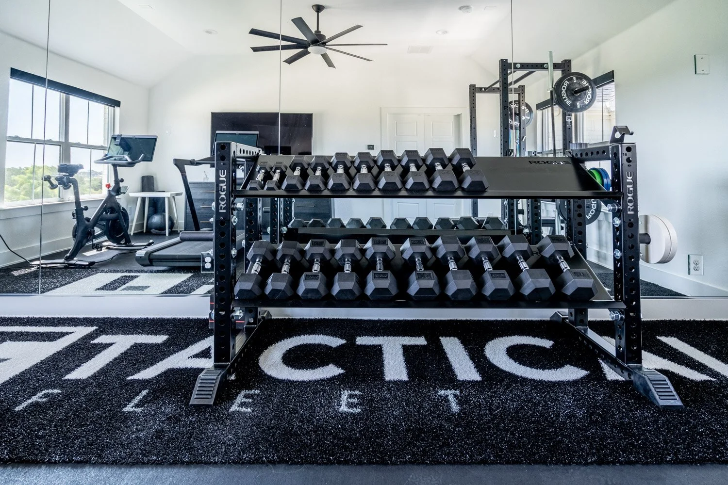 Spacious home gym with Rogue dumbbell rack, treadmill, stationary bike, mirrored wall, and bright natural lighting