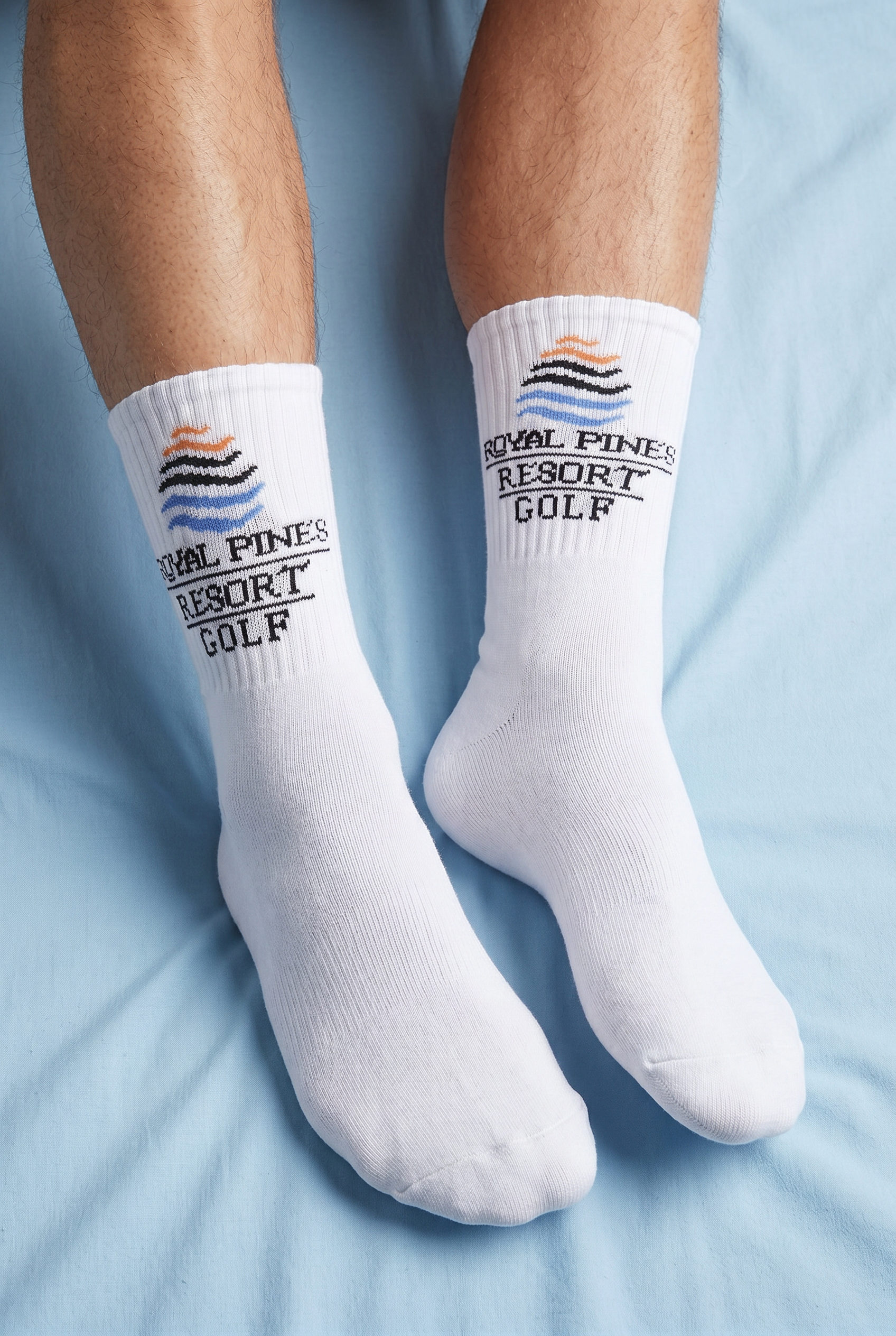 Close-up of a person's legs wearing white Socks with a logo that reads 'Royal Pines Resort Golf' and a stylized wave emblem, against a light blue background.
