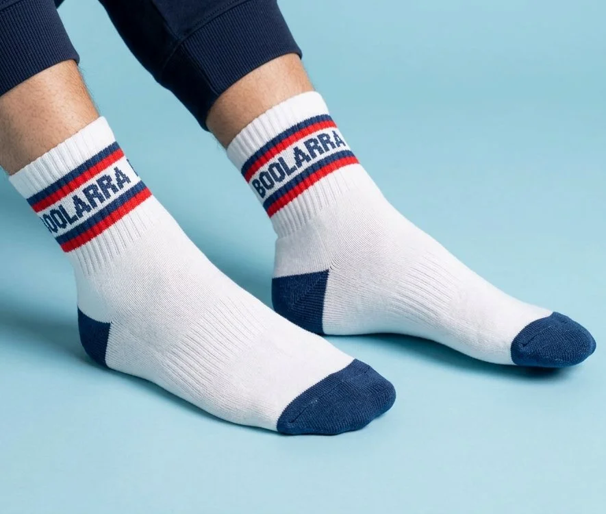Legs of person with custom white sports quarter socks which have navy heel and toe, navy and red stripes, and branded with text 'BOOLARRA' knitted into the socks