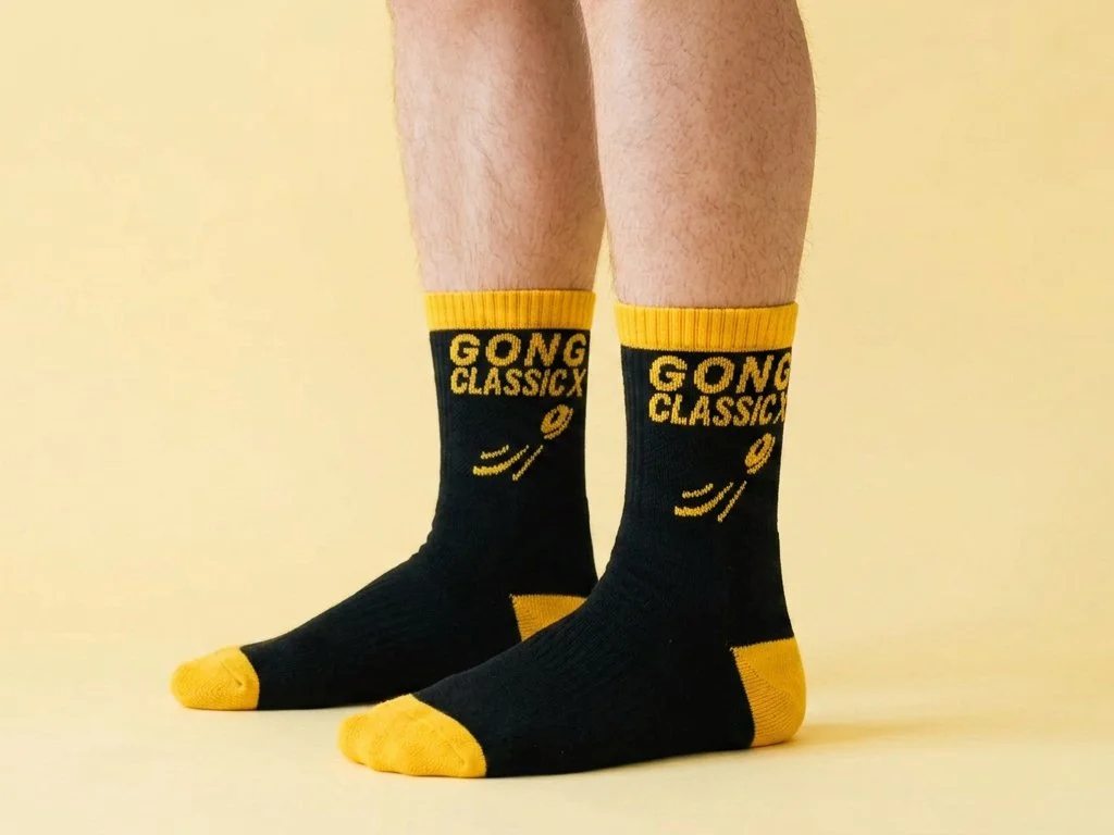 Legs of person wearing black sports quarter socks with yellow heel, toe and welt and text 'GONG CLASSICS' knitted into sock. Person against a yellow background