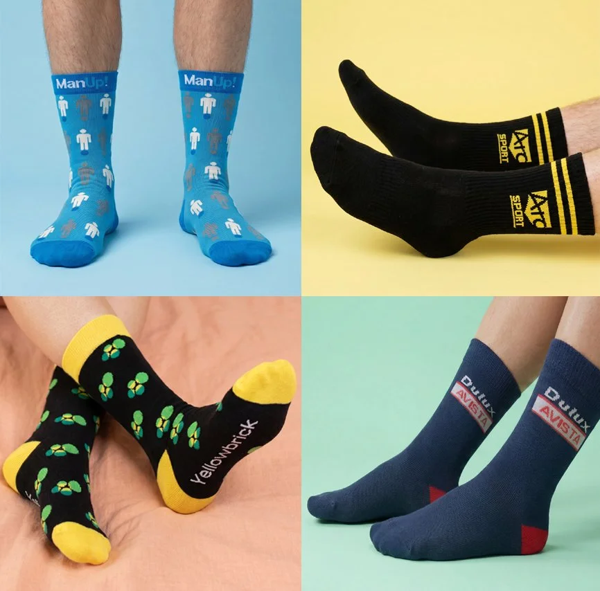 Four pairs of socks with different cudesigns and colors: blue socks with white and brown human figures and grey hands, black socks with yellow accents and sport logo, black socks with green and yellow clovers, and blue socks with red and white logos.