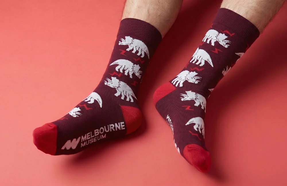 Person wearing maroon socks with white and red dinosaur designs and 'Melbourne Museum' logo on the toes, against a pink background.