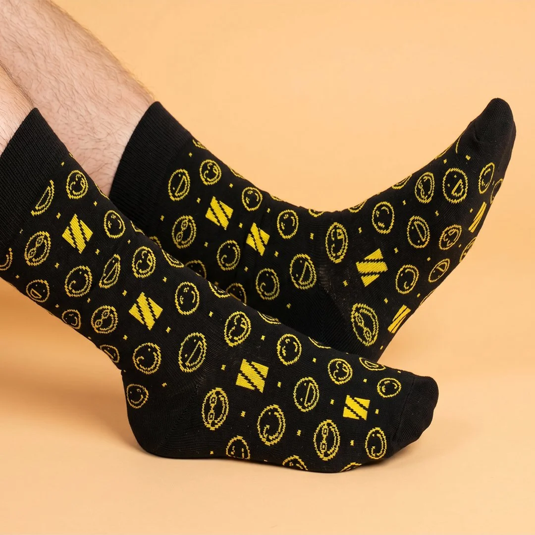 Person wearing black socks with yellow smiley faces, credit card icons, and other symbols, against a beige background.