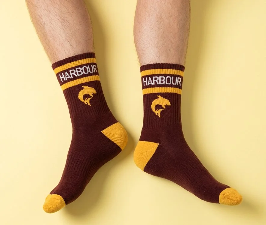 Legs of person lying down and wearing custom sports quarter socks in maroon and gold with shark and text 'HARBOUR' knitted into socks