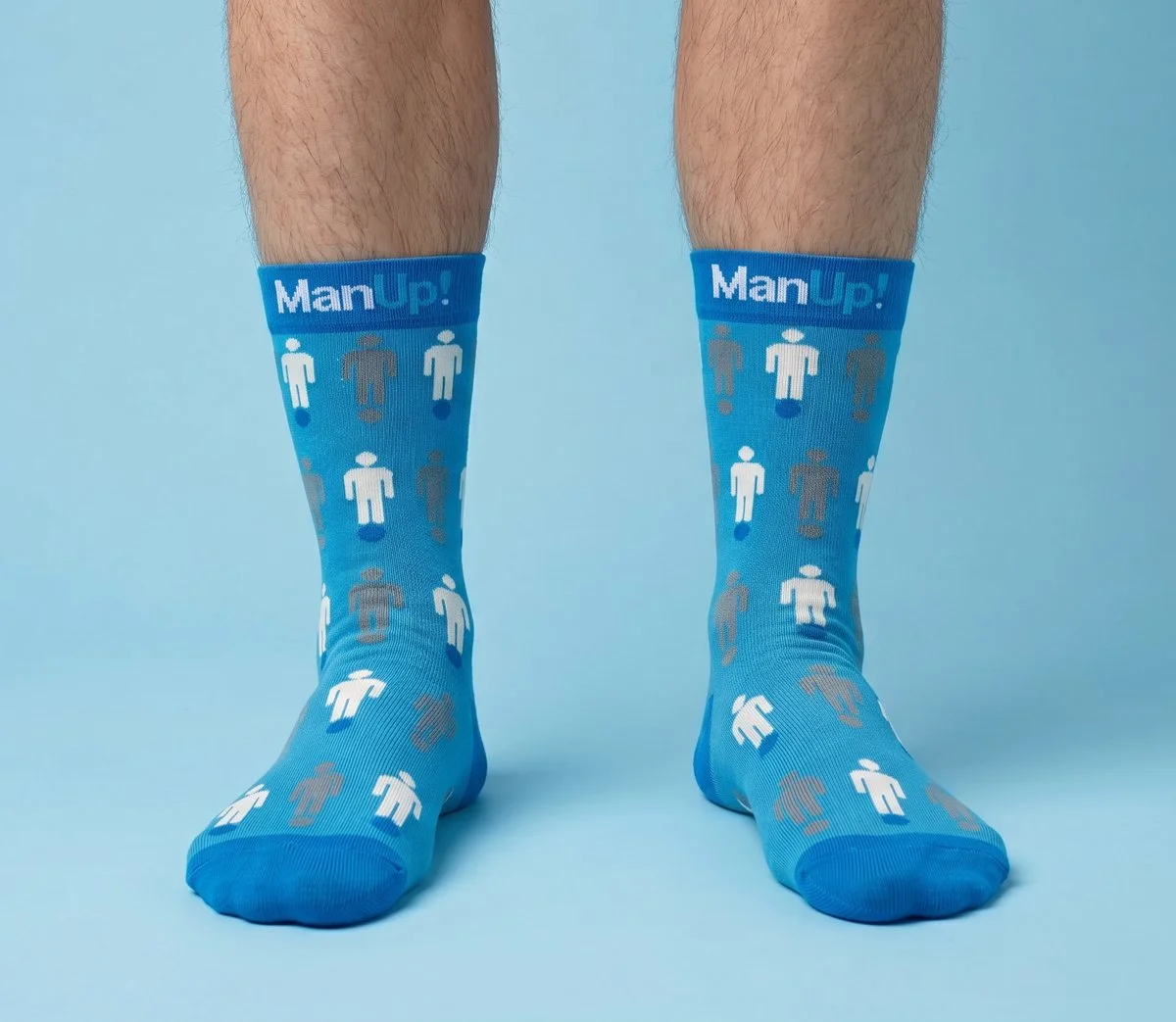Pair of blue socks with white and beige human figures knitted into the sock and the phrase 'Man Up!' at the top, worn on hairy legs against a light blue background.