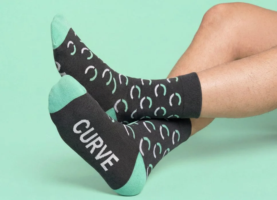 A person wearing black and teal socks with a pattern of white and teal circles on a light green background, and the word 'CURE' written on the sole of the sock.
