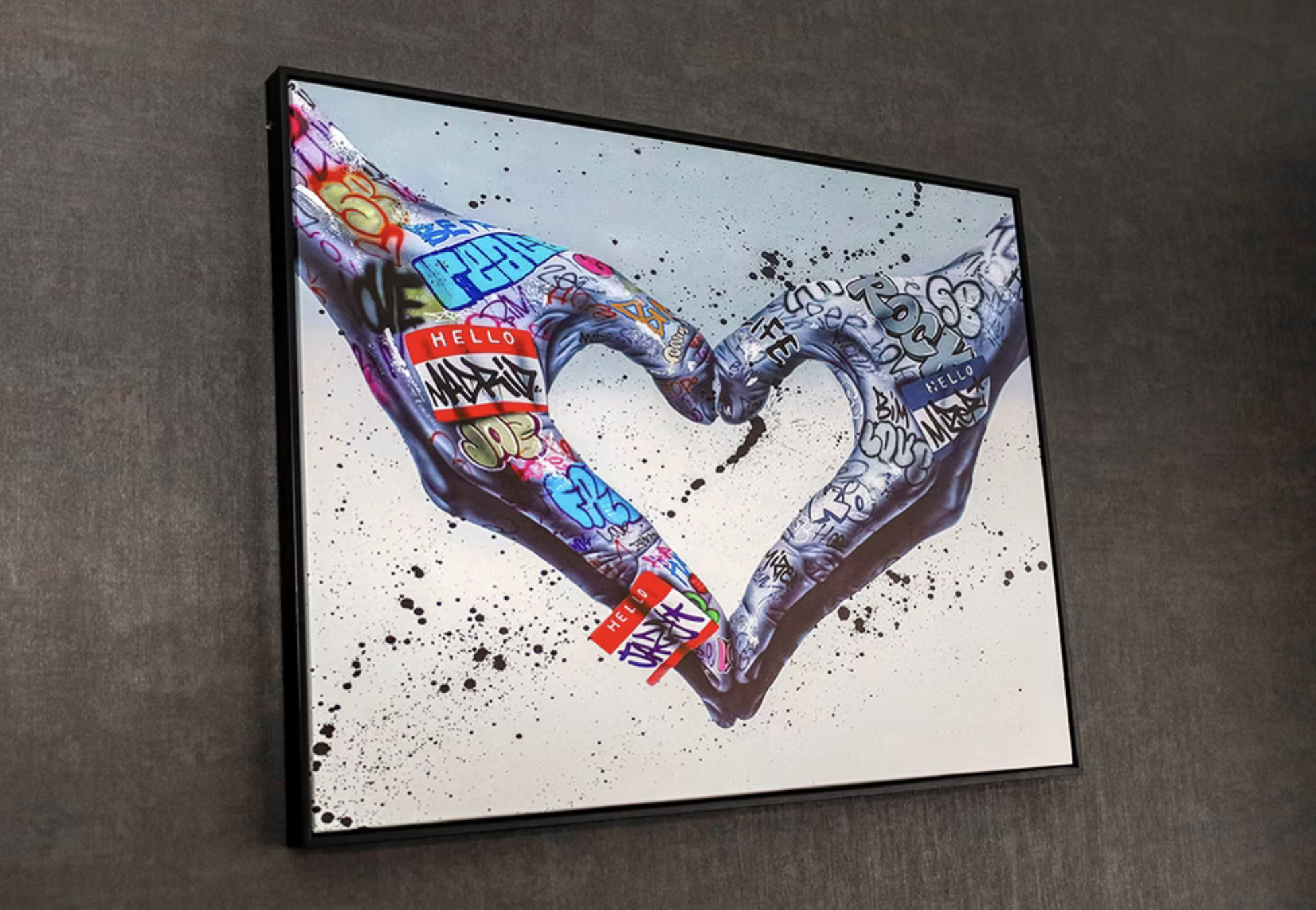 A framed digital artwork of two hands forming a heart shape with graffiti-style text and colors, on a gray wall with black splatter paint.