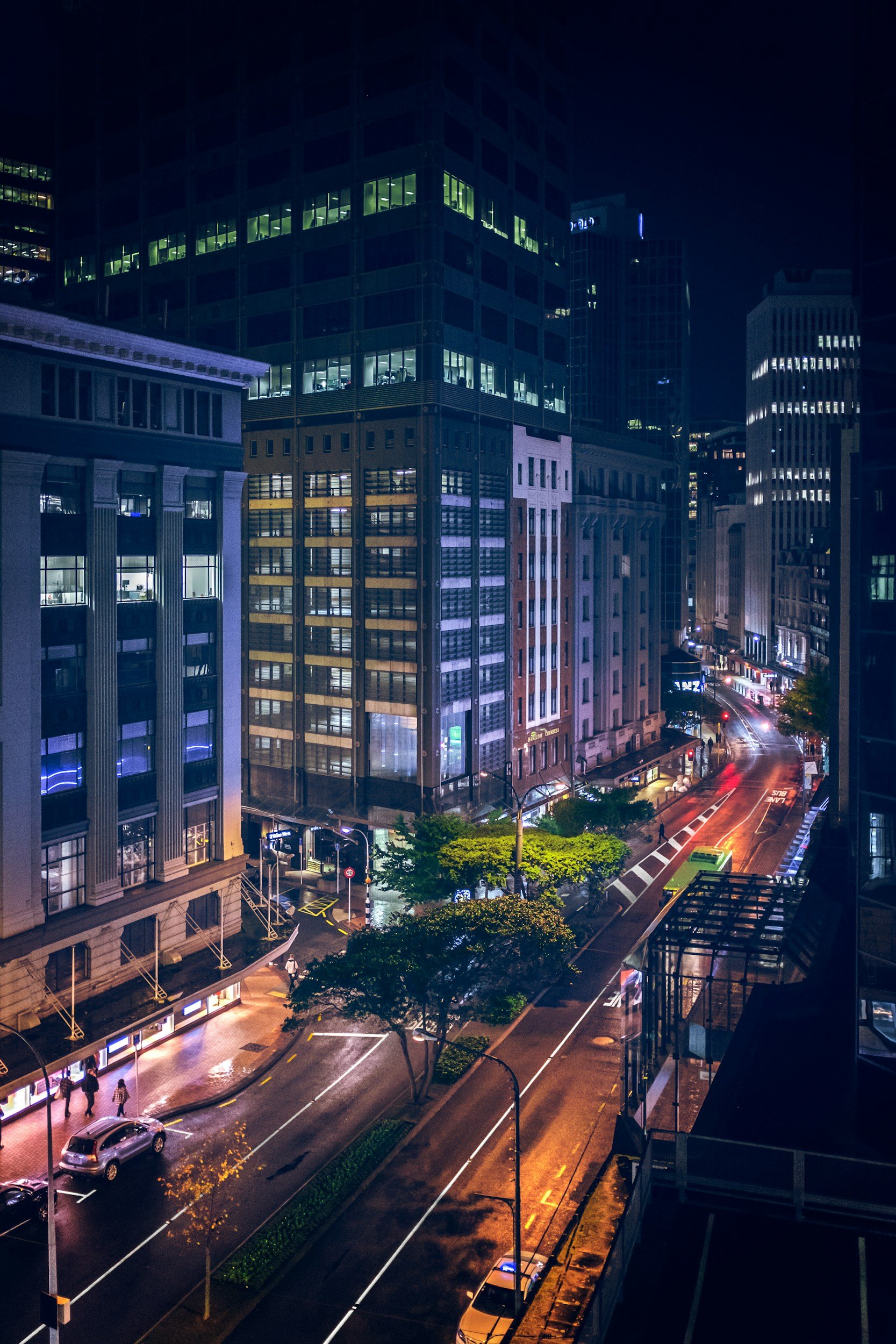 Night view of tall office buildings with illuminated windows, a city street with some cars, trees, and pedestrians, and a wet road reflecting streetlights.