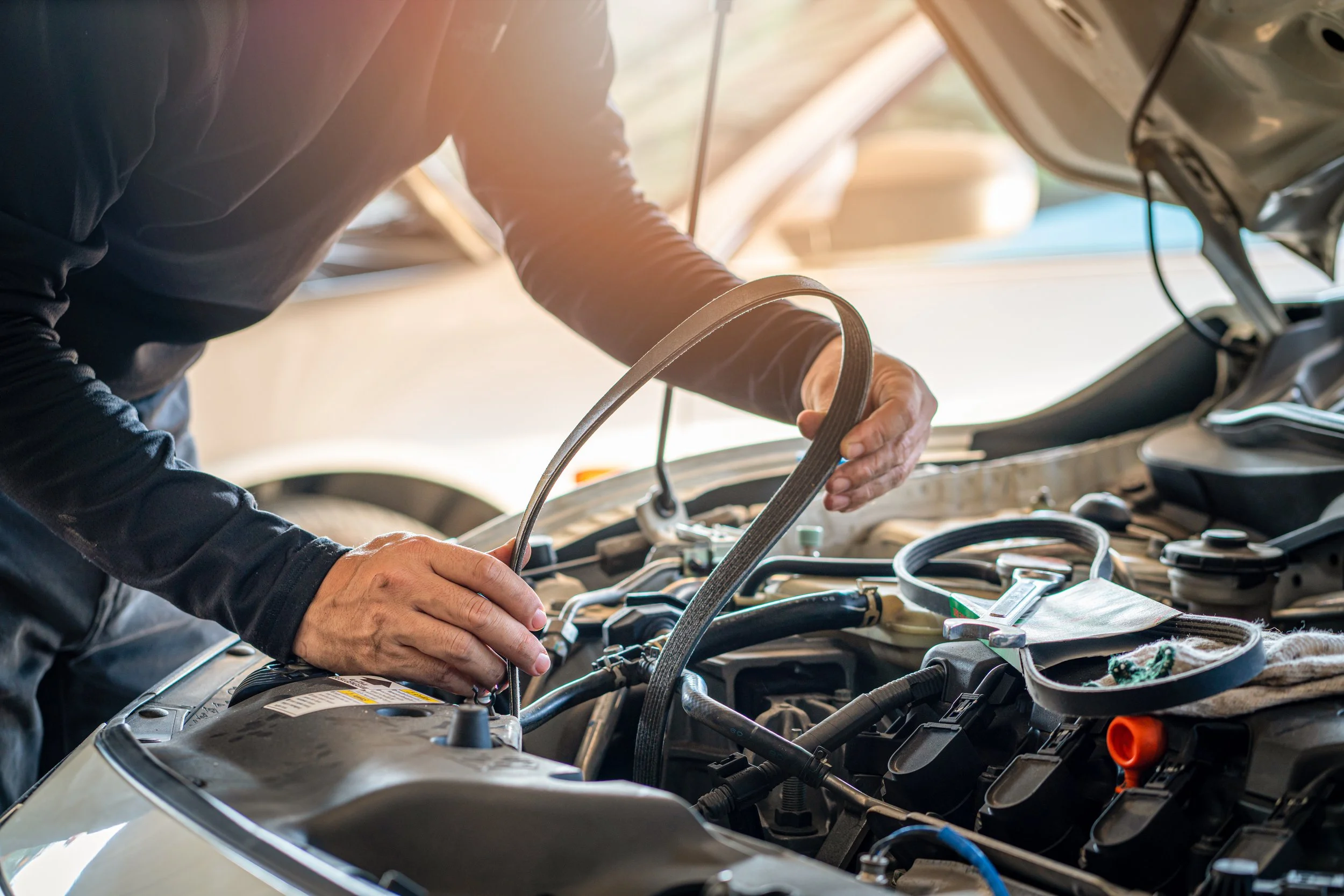 Person working on car engine with hood open, holding a belt or hose, in a well-lit garage or workshop.