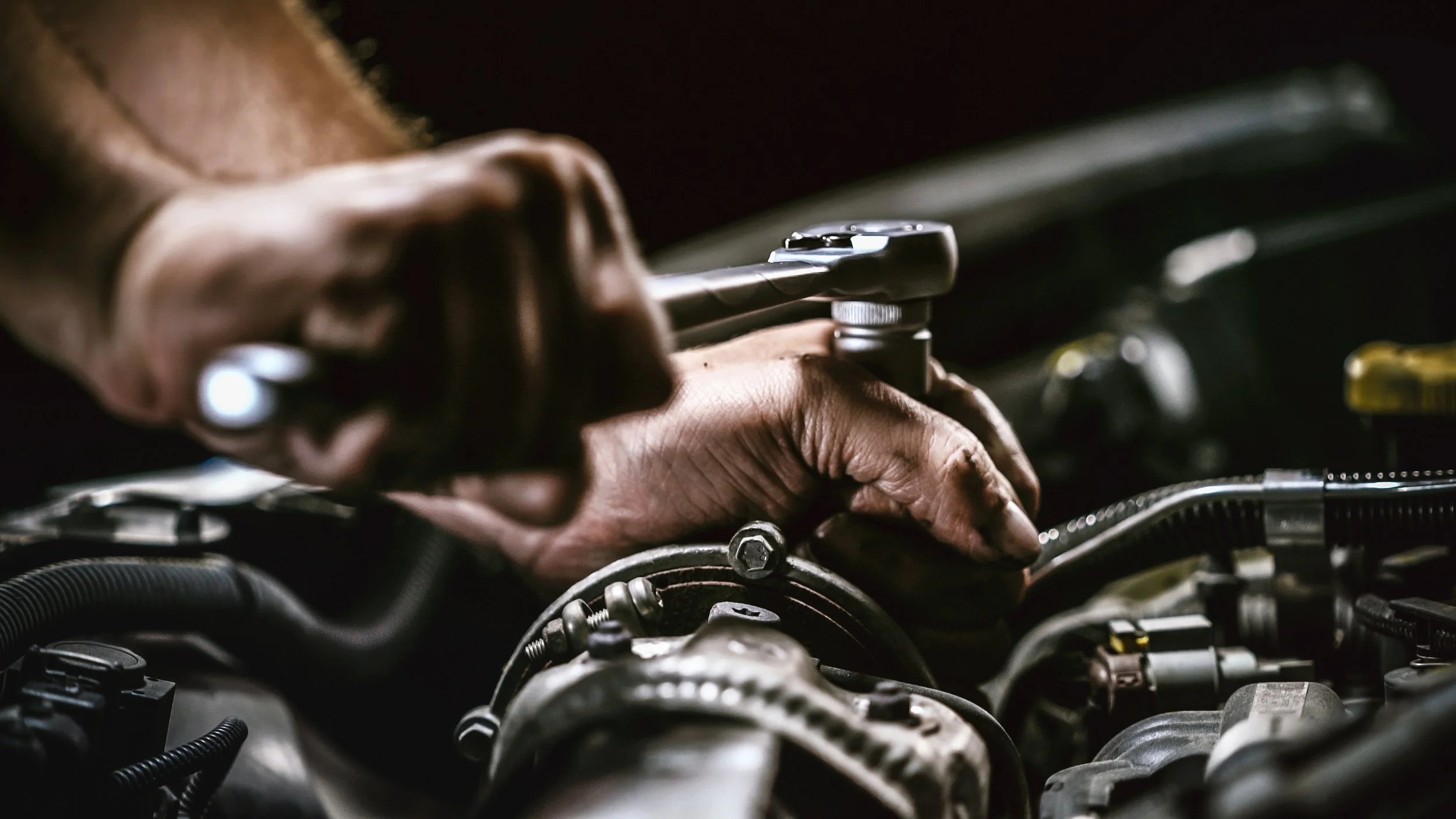 A mechanic's hand holds a ratchet wrench while working on a car engine in a dimly lit space.