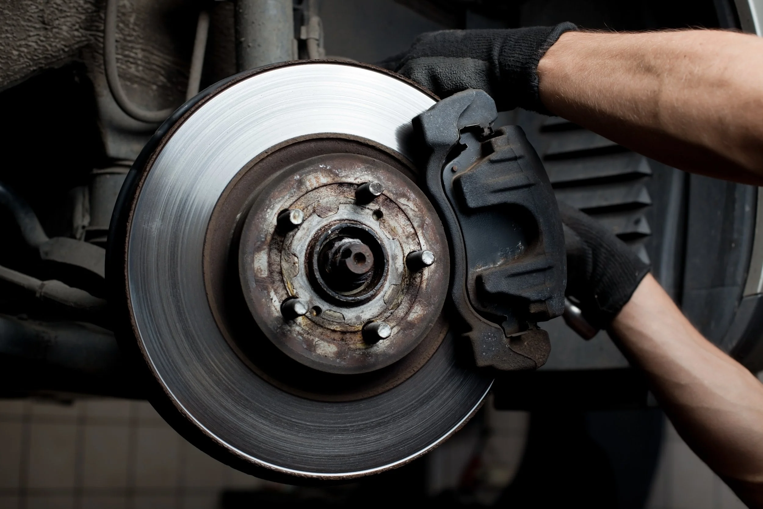 A person wearing black gloves is working on a car's brake disc and caliper, which appears rusty and worn, with a dark background.