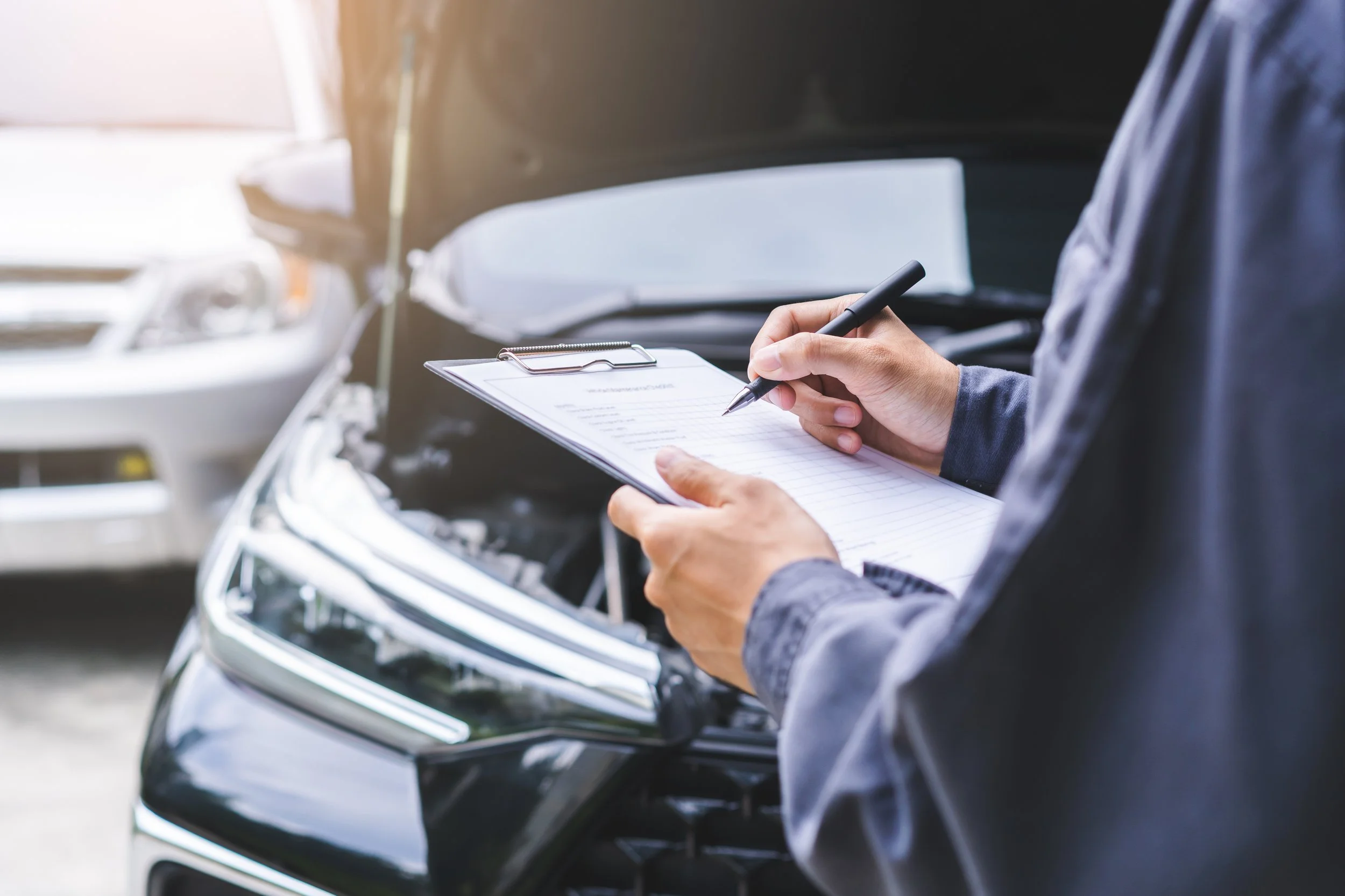 A person holding a clipboard and writing with a pen near a car with its hood open, in an outdoor setting.