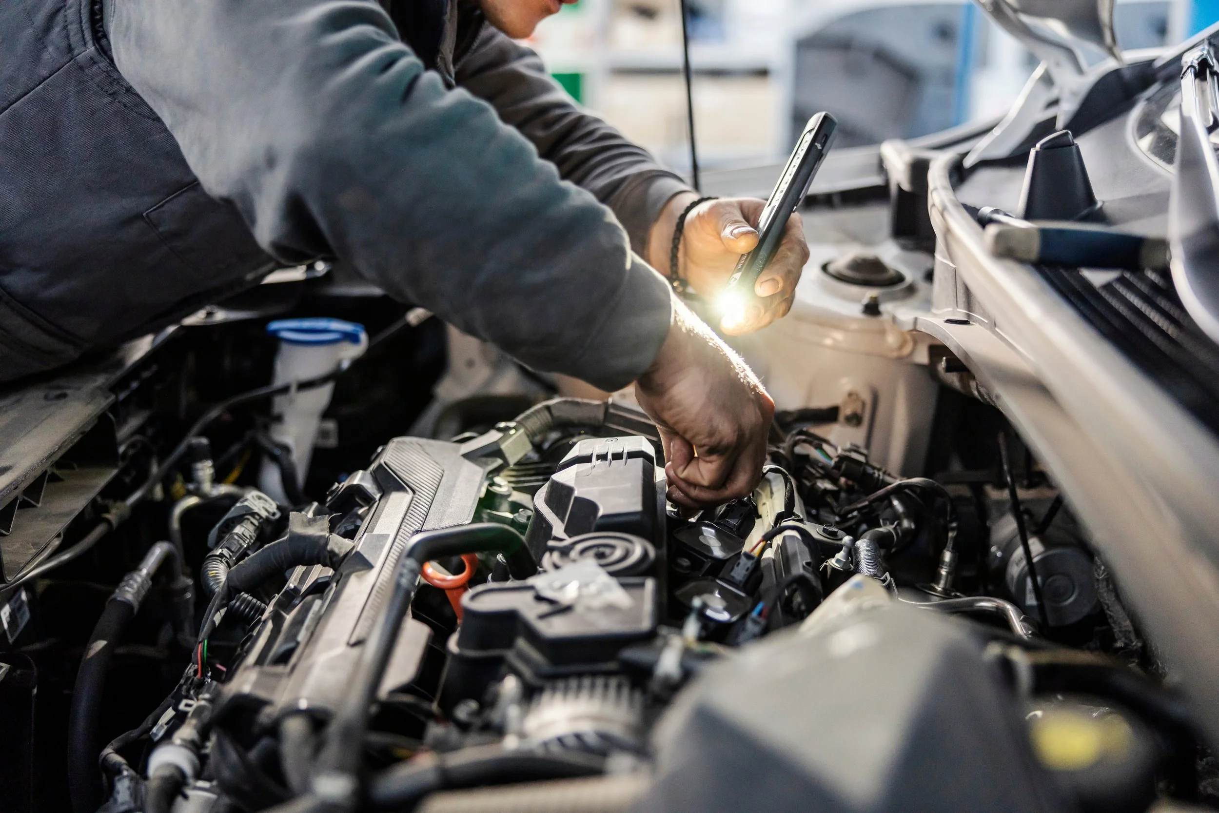 A person working on a car engine, holding a smartphone in one hand and a tool in the other, with various engine parts and wires visible.