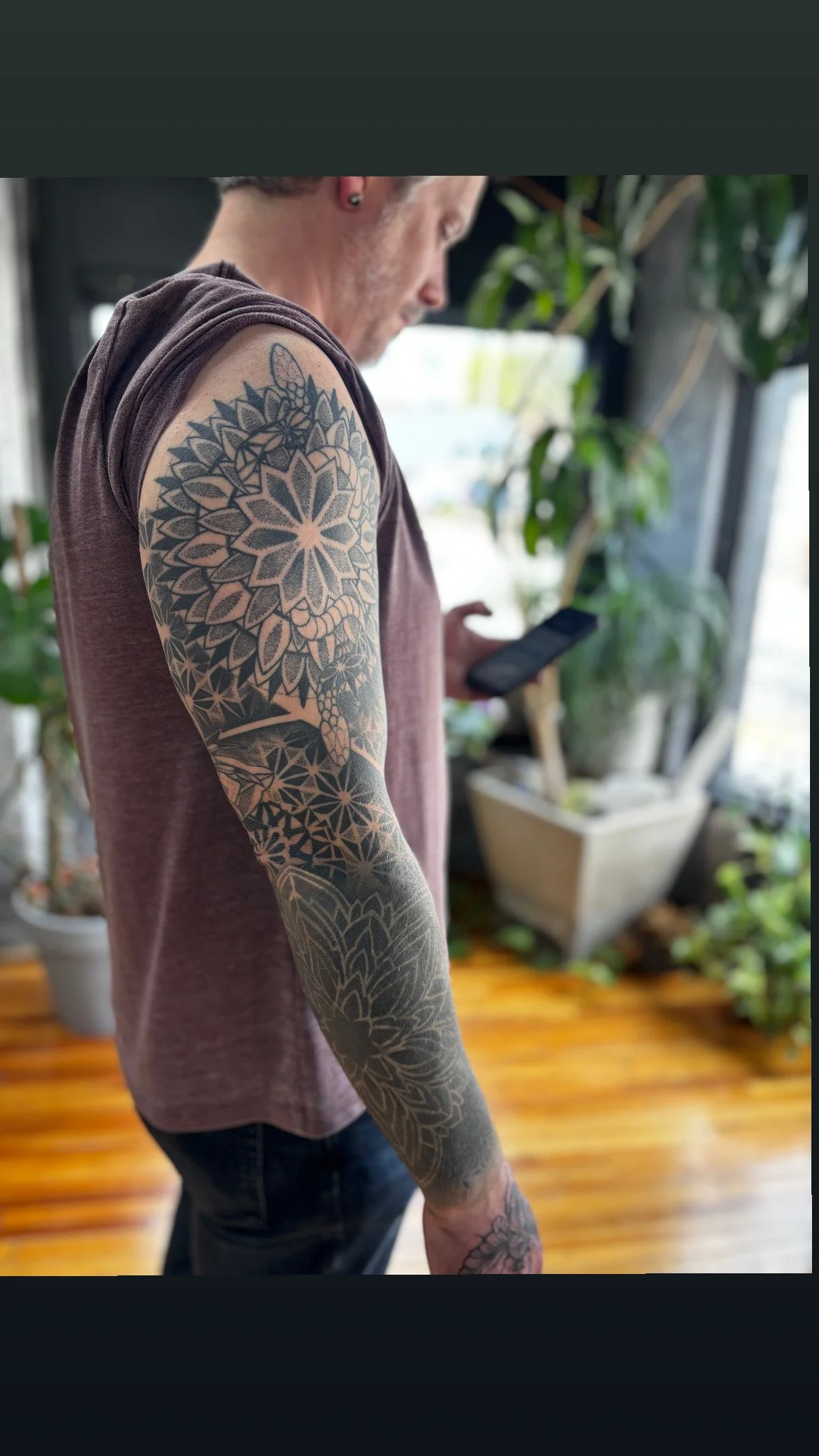 A man with tattoos on his arm, wearing a sleeveless shirt, looking at his phone in a room with plants and wooden flooring.