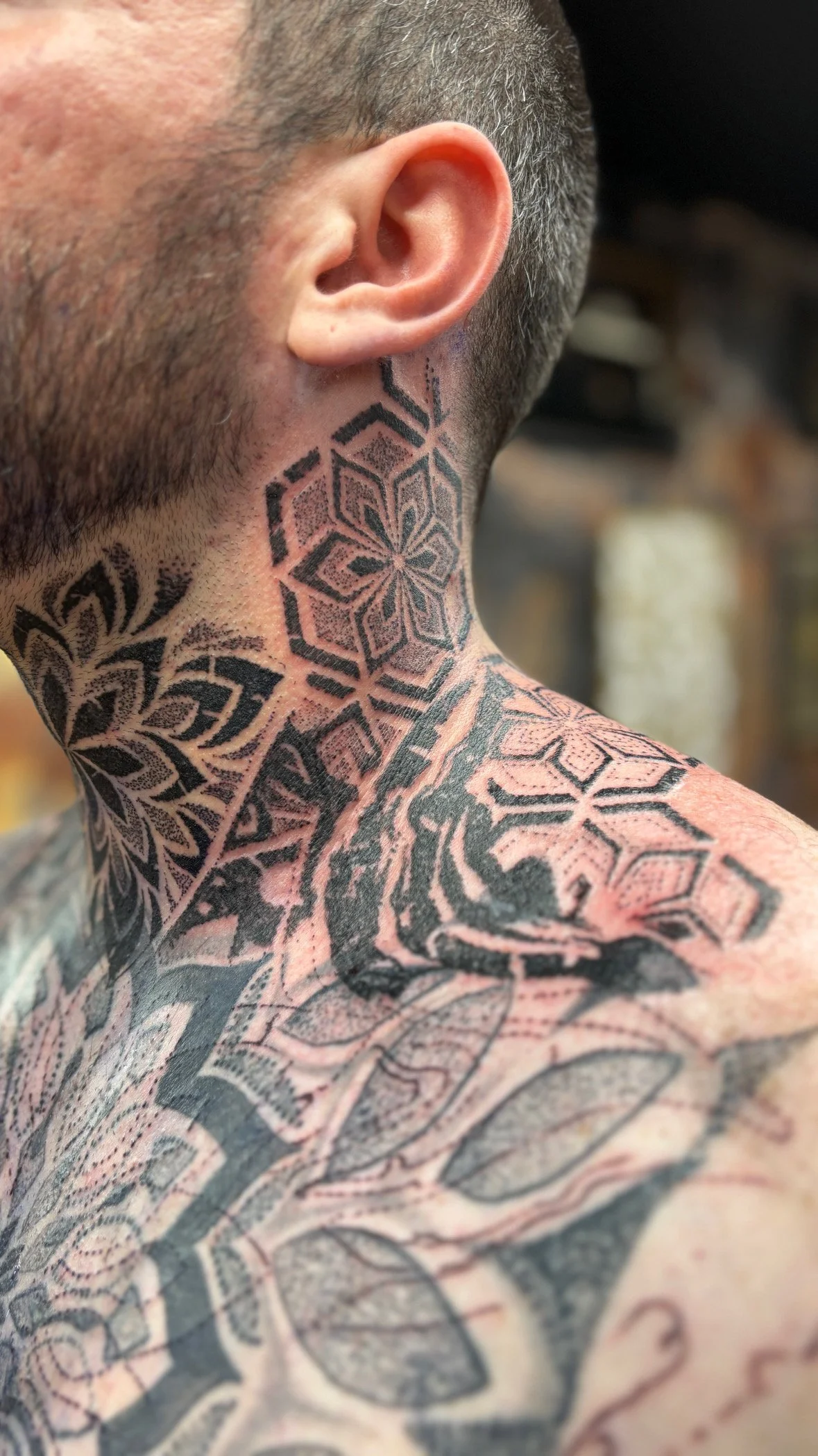 Close-up of a man's neck and shoulder covered in detailed black tattoos with geometric patterns.