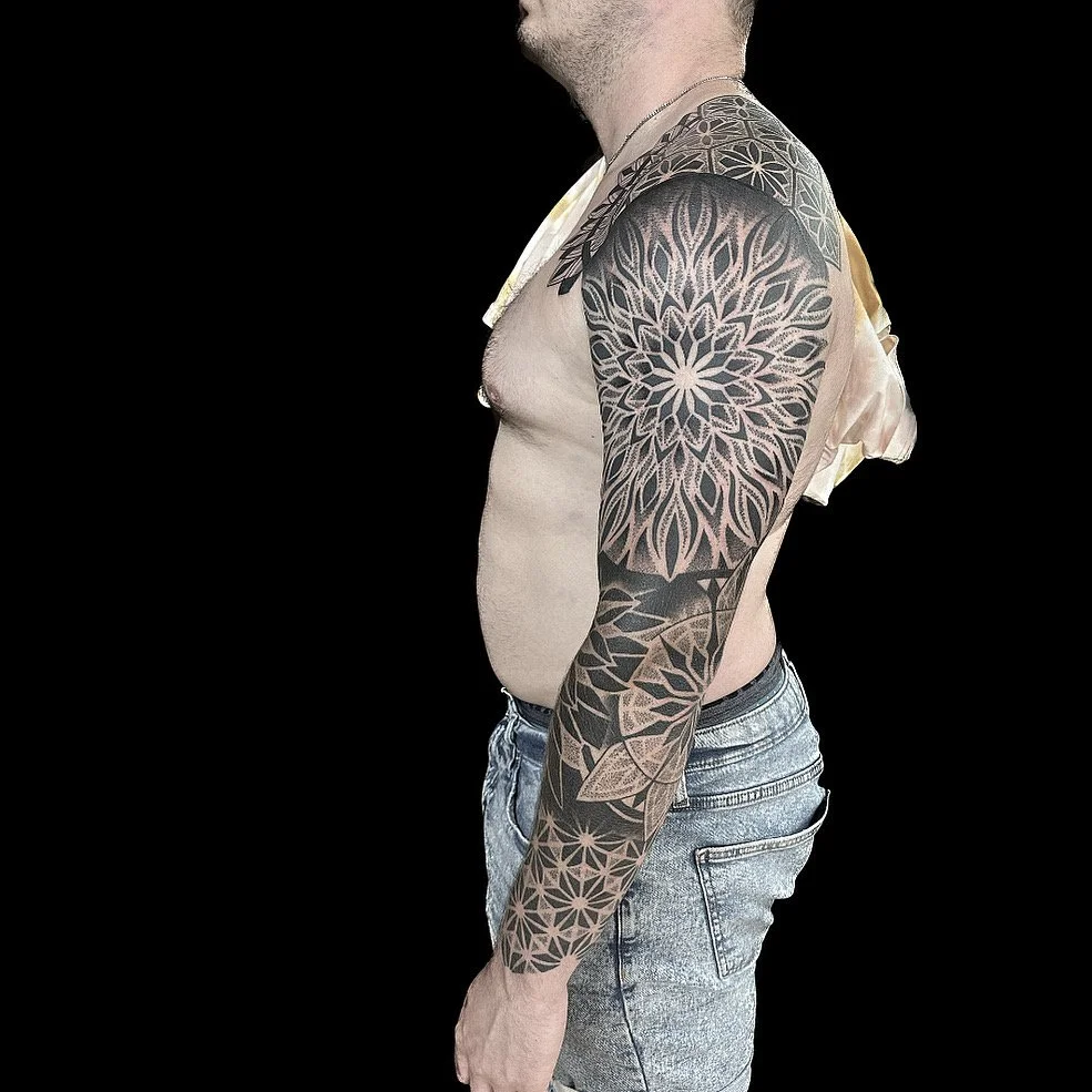 A man with a large black and gray geometric and floral tattoo sleeve on his right arm, shown in profile against a black background, wearing gray denim shorts and no shirt.