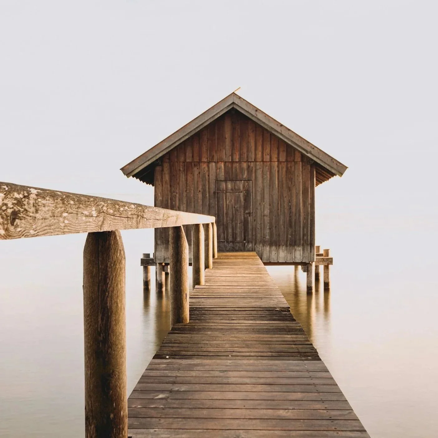 A wooden dock leading to a small wooden hut over calm water, with a simple railing on the left side and a cloudy sky in the background.