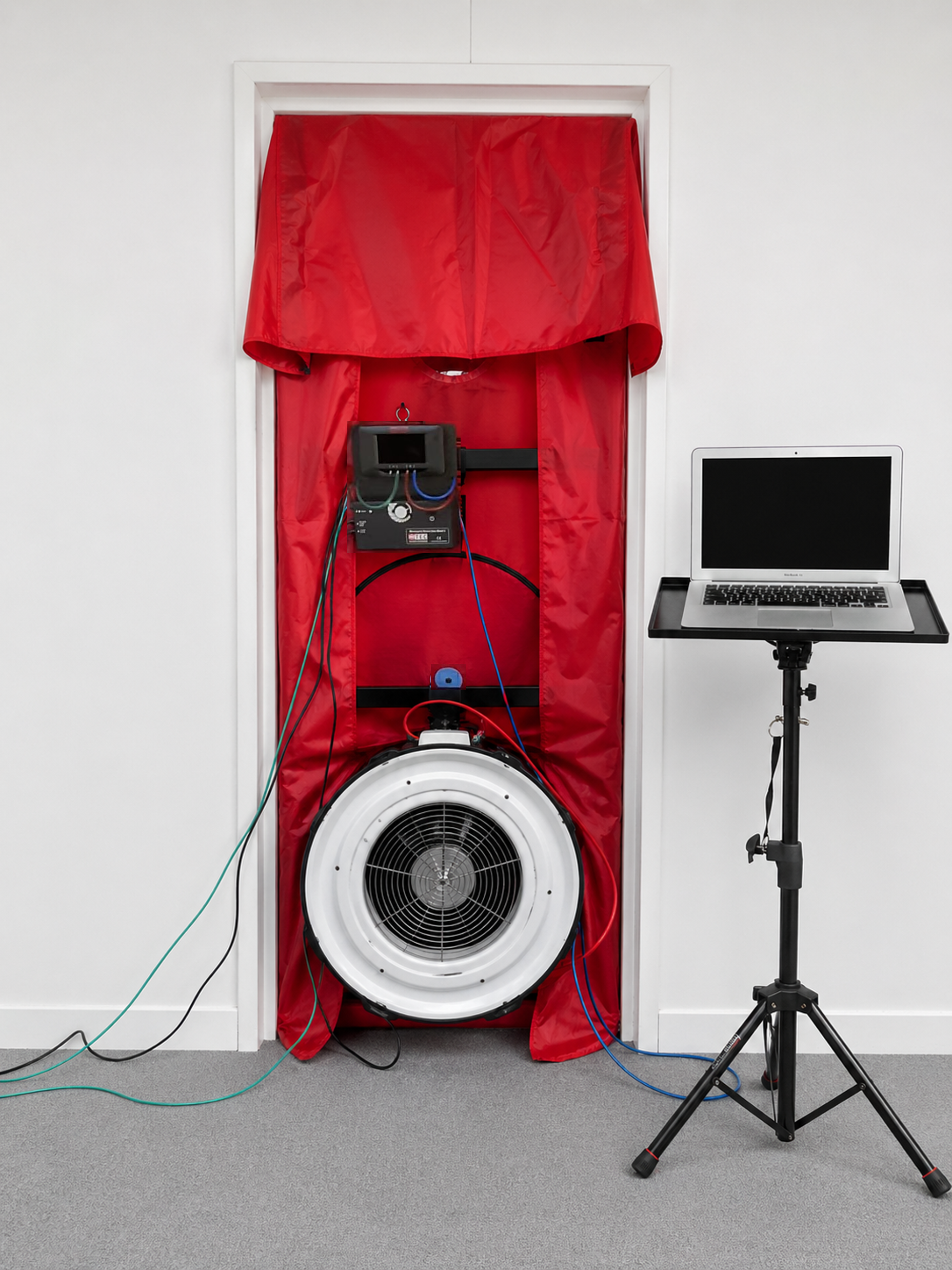 A red couch with a large speaker in front of it, connected with multiple colored wires, inside a white room, with a laptop on a stand to the right.