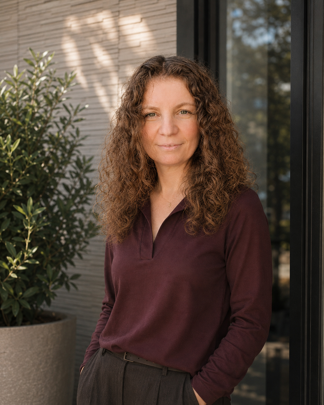 A woman with curly brown hair and blue eyes stands outdoors near a modern building with large windows and a potted plant, wearing a maroon long-sleeve shirt and dark pants.