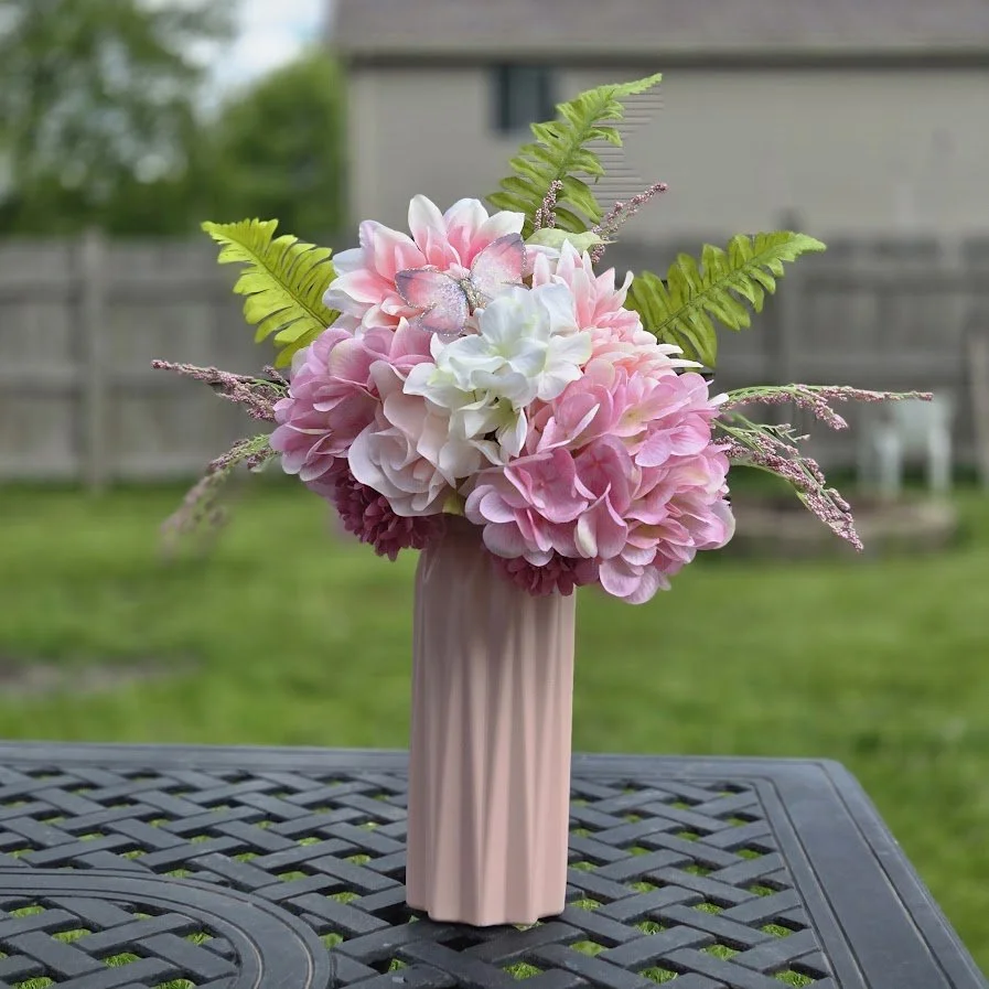 A pink ceramic vase with a textured surface holds a bouquet of pink and white hydrangeas and greenery, placed outdoors on a black metal table.