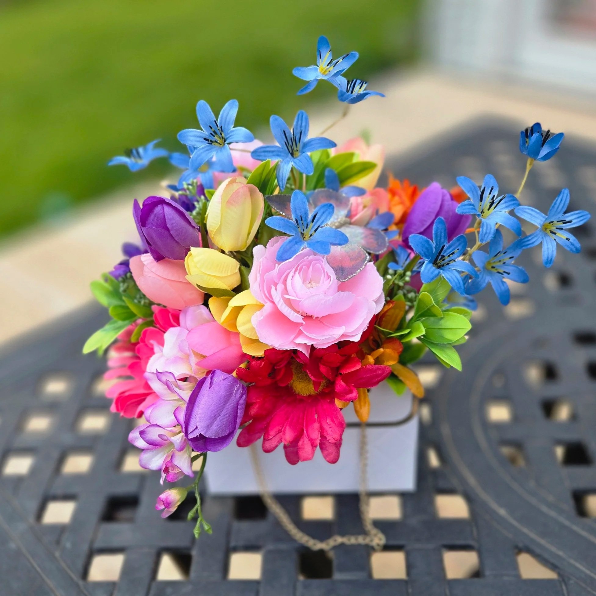 A close-up of a colorful artificial flower bouquet with blue, purple, pink, yellow, and orange flowers, with a chain necklace hanging over the flowers, set against a blurred outdoor background.